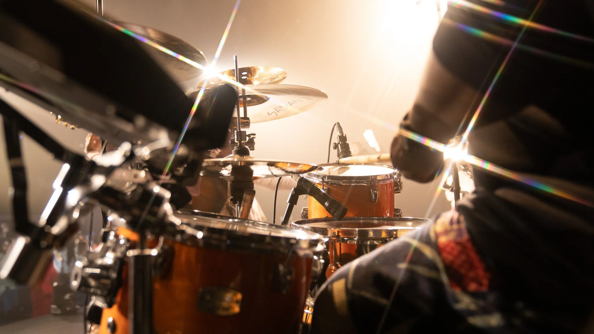 Close-up of a drum set being played on stage with bright stage lighting reflecting off the cymbals and drum shells.