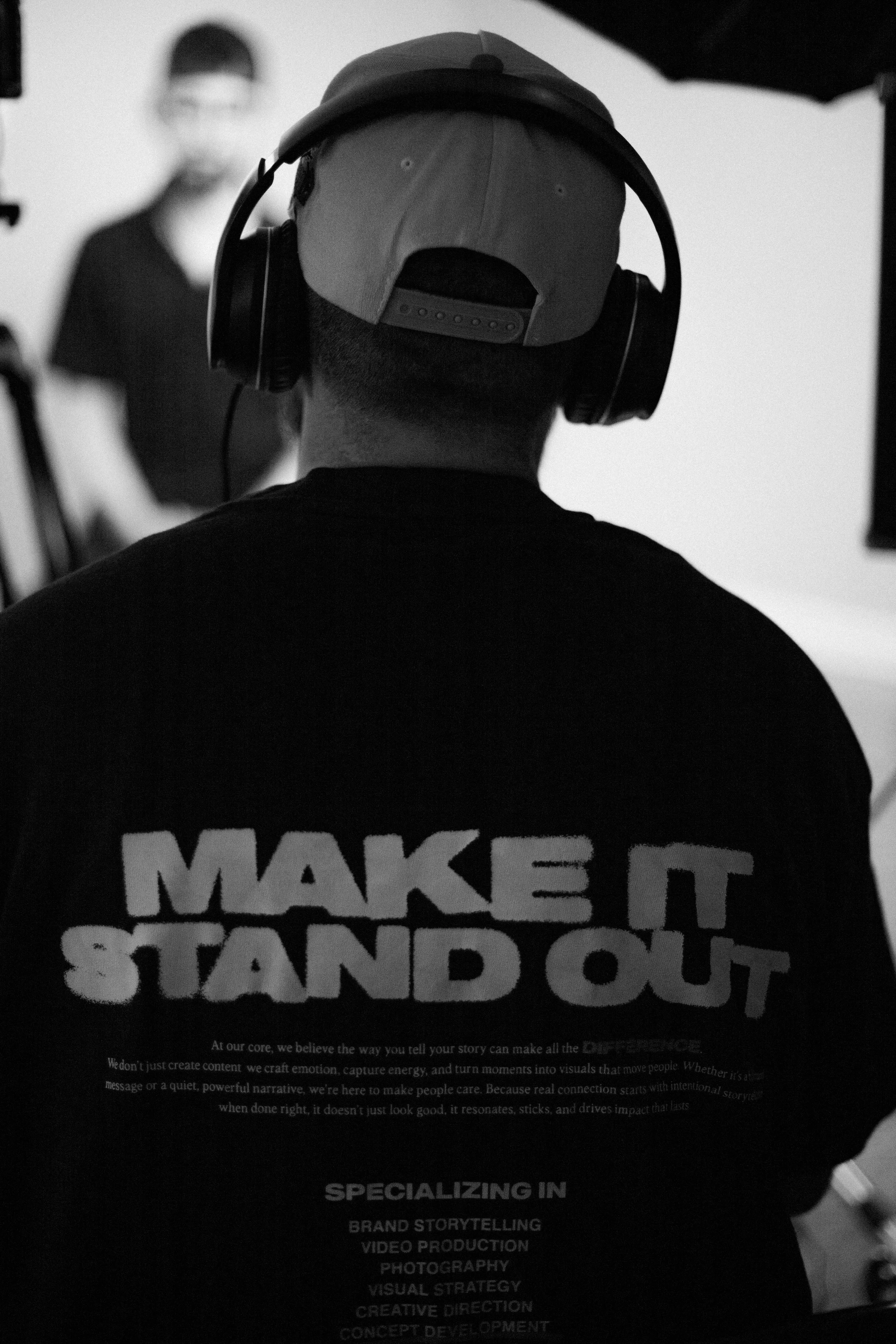Back of a person wearing a cap and headphones, with the shirt reading 'MAKE IT STAND OUT' and additional text about brand storytelling and video production.