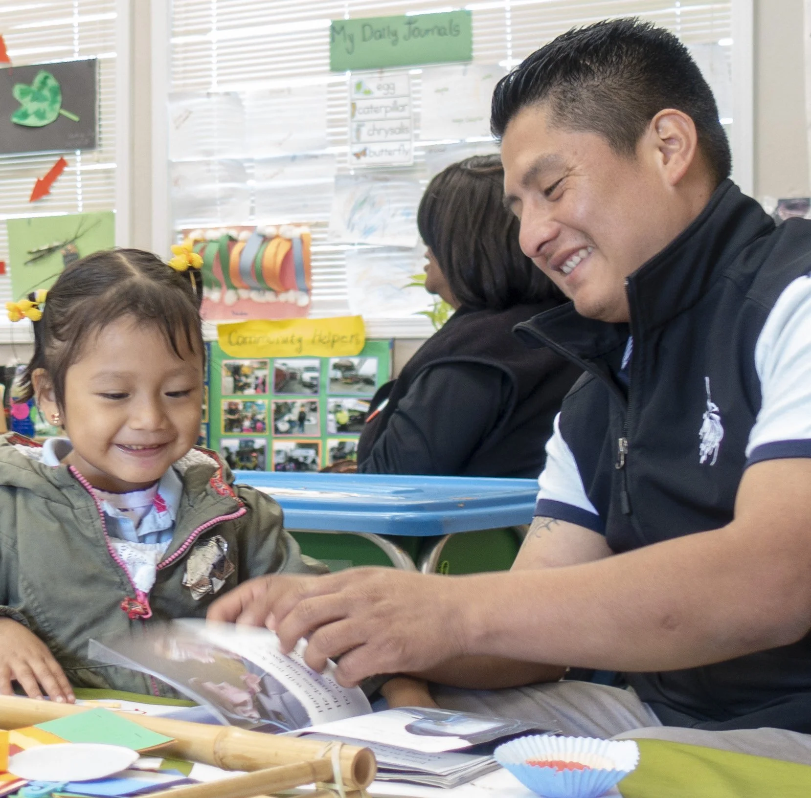 A young girl with a yellow hair bow, sitting at a table, smiling at an adult man. The setting is a classroom with colorful educational posters on the window and a sign that says 'My Daily Journals.' The man is smiling and engaging with the girl, with various craft supplies and books on the table.