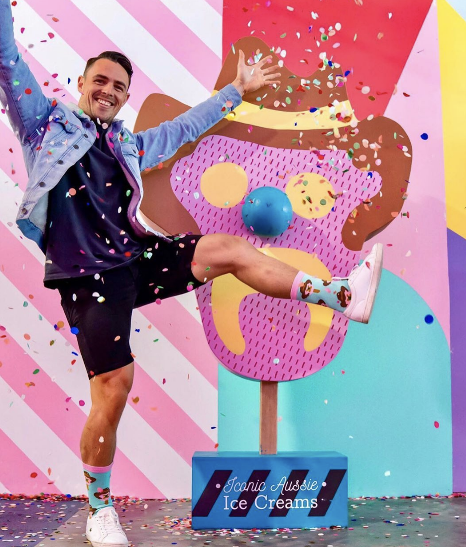 A man celebrating with a big smile, kicking one leg up, in front of a colorful ice cream-themed backdrop. He is surrounded by falling confetti. He wears a blue jacket, black shorts, white sneakers, and colorful socks with cartoon characters.