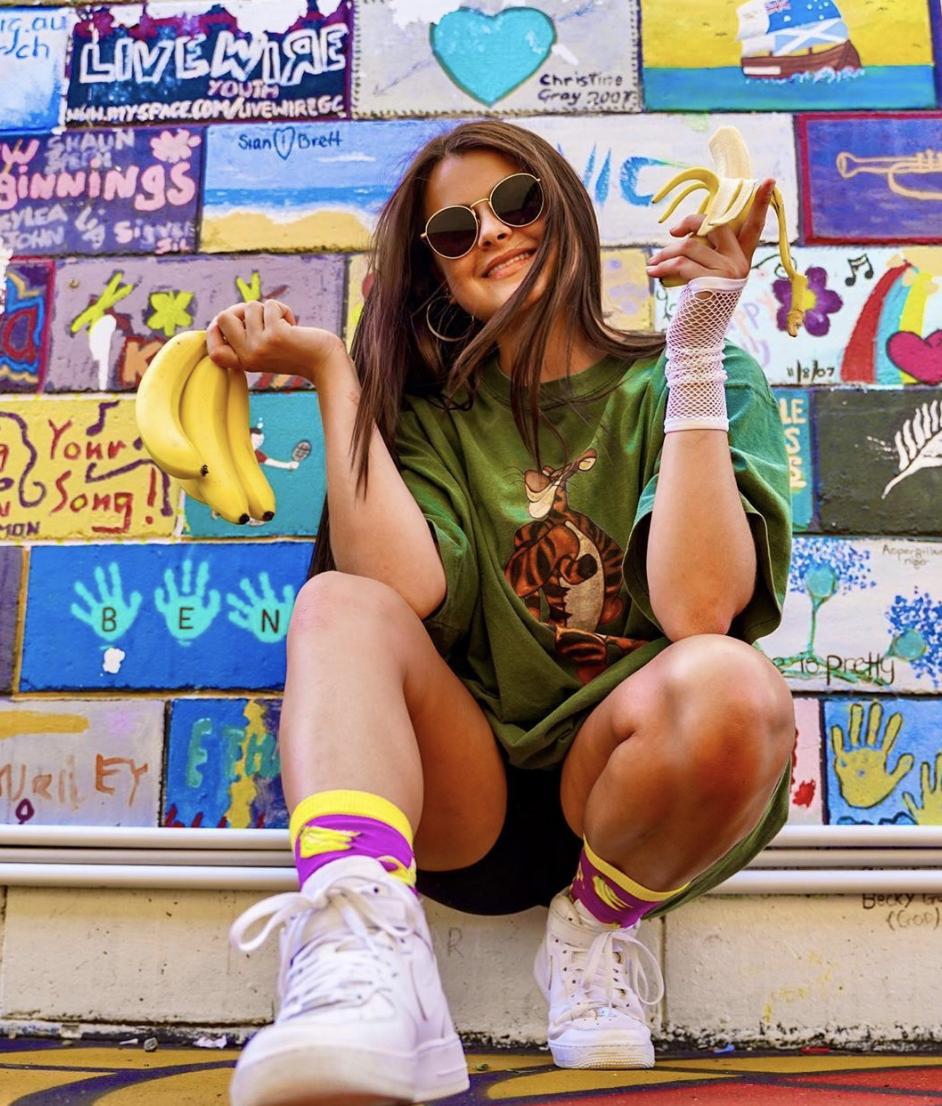 A young woman with long brown hair, wearing round sunglasses, a green T-shirt with a tiger graphic, fishnet gloves, and white sneakers, sitting on a step in front of a colorful, painted brick wall. She is holding a bunch of bananas in her right hand and a peeled banana in her left hand, smiling at the camera. The wall behind her features various vibrant murals, drawings, and writings.