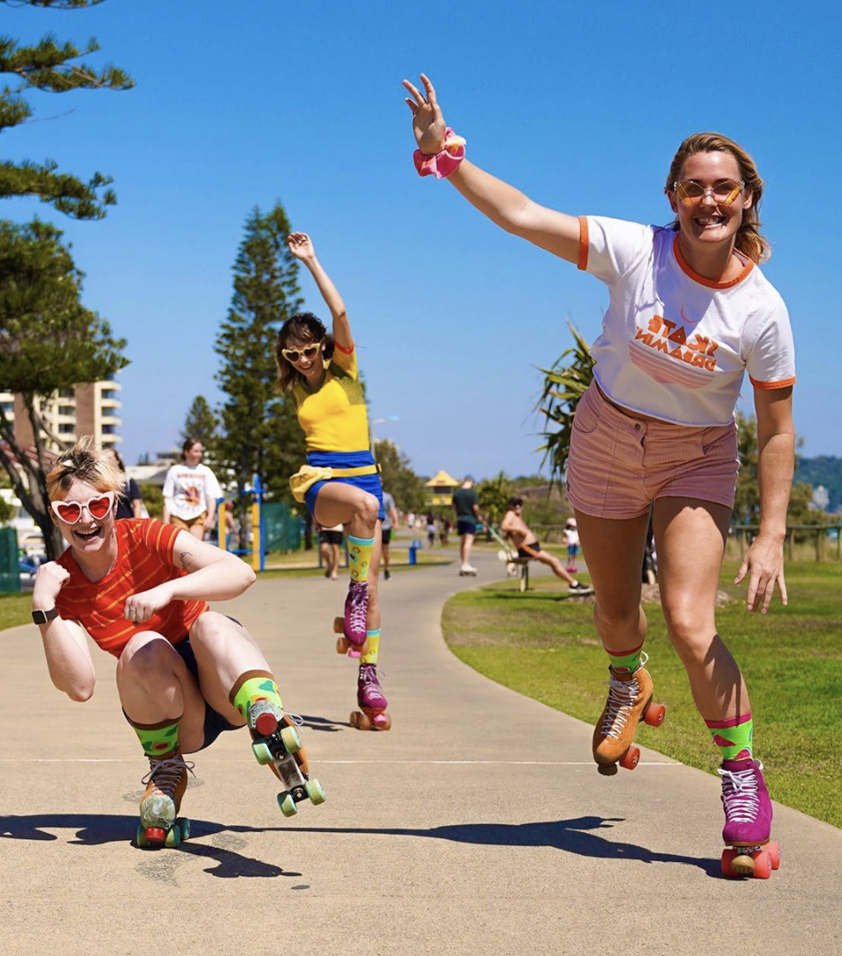 Four people roller skating in a park on a sunny day, smiling and waving. They are wearing colorful clothes and accessories, with some sporting heart-shaped sunglasses.