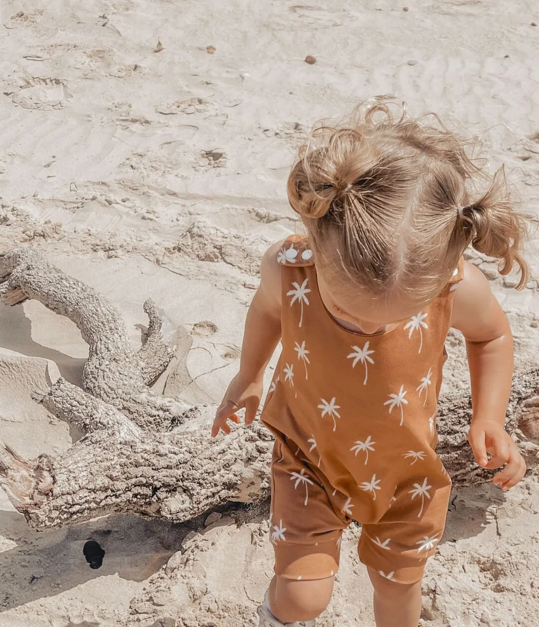 A young girl with curly blonde hair in pigtails explores a sandy beach near a piece of driftwood wearing a handmade Little Spumkins romper