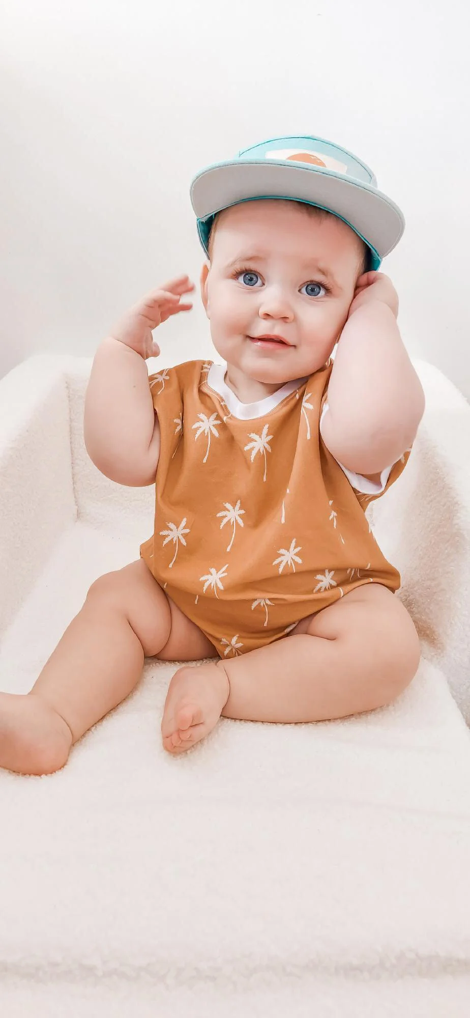 A young child with blue eyes, wearing a tan shirt with white palm tree prints and a gray baseball cap, sitting on a white blanket and touching their head.