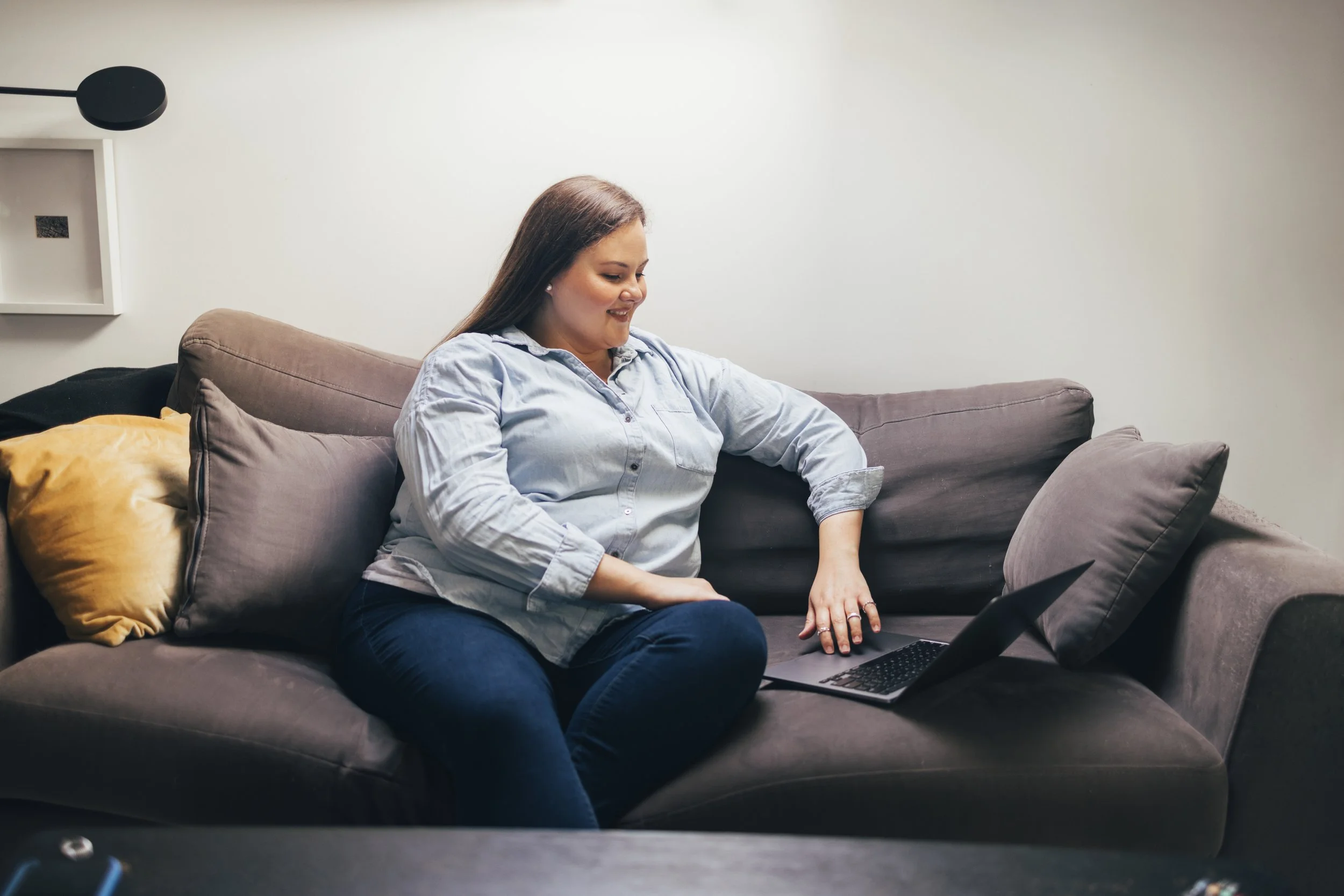 A smiling woman with brown hair wearing a light blue shirt sitting on a gray couch in a living room, working on a laptop.