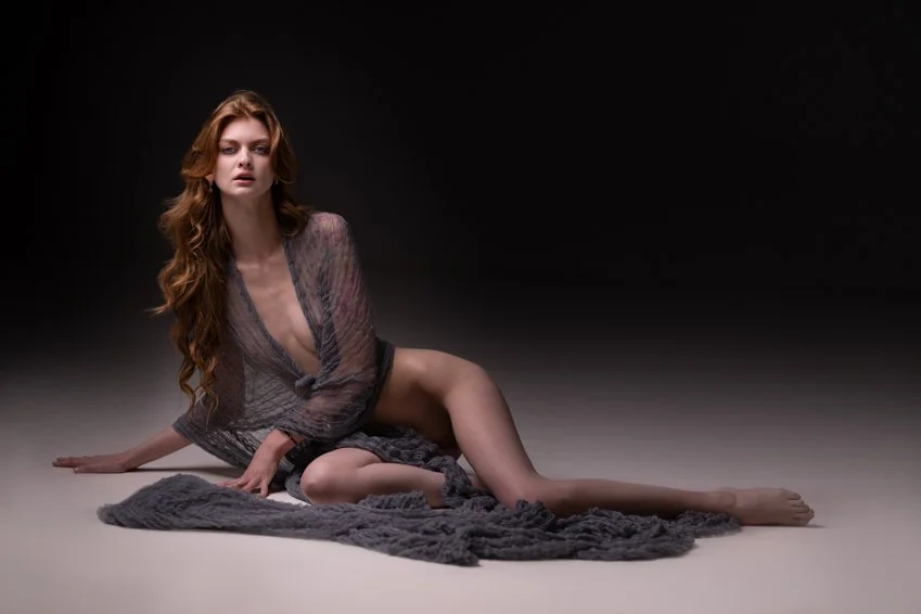 A woman with long, wavy red hair is sitting on the floor with a dark background. She is dressed in a sheer, loose dress and lying partially on a textured gray blanket.