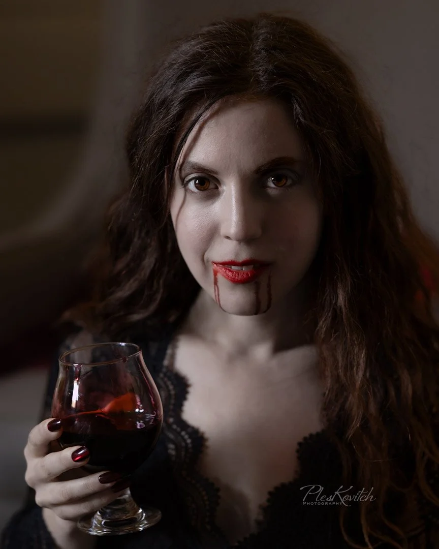 A woman with long, dark wavy hair and pale skin holding a glass of red wine. She has blood on her lips and dripping from her mouth, and she is wearing dark makeup and a black lace top.