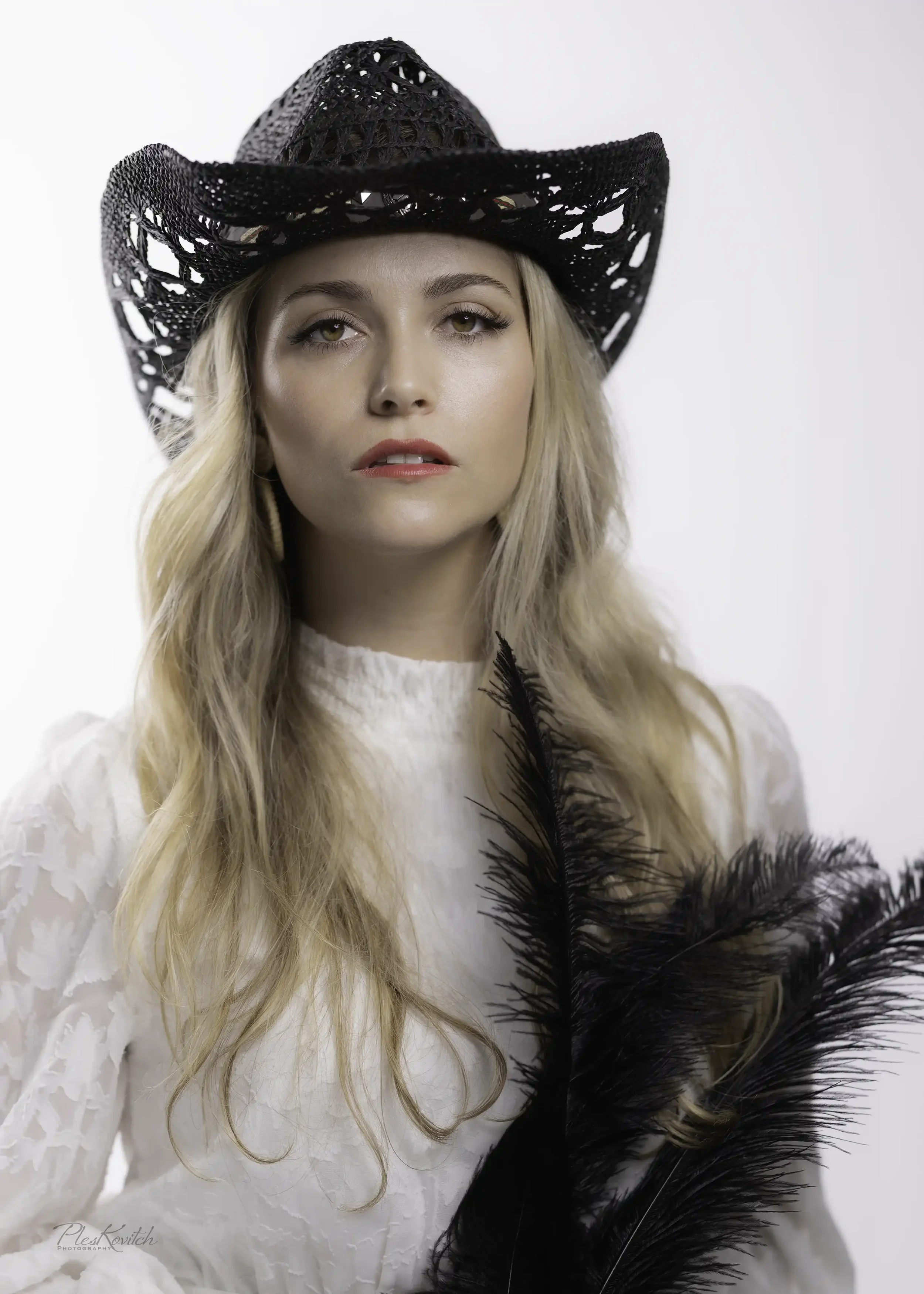 A woman with long blonde hair wearing a black woven cowboy hat, a white lace dress, and holding black feathers.
