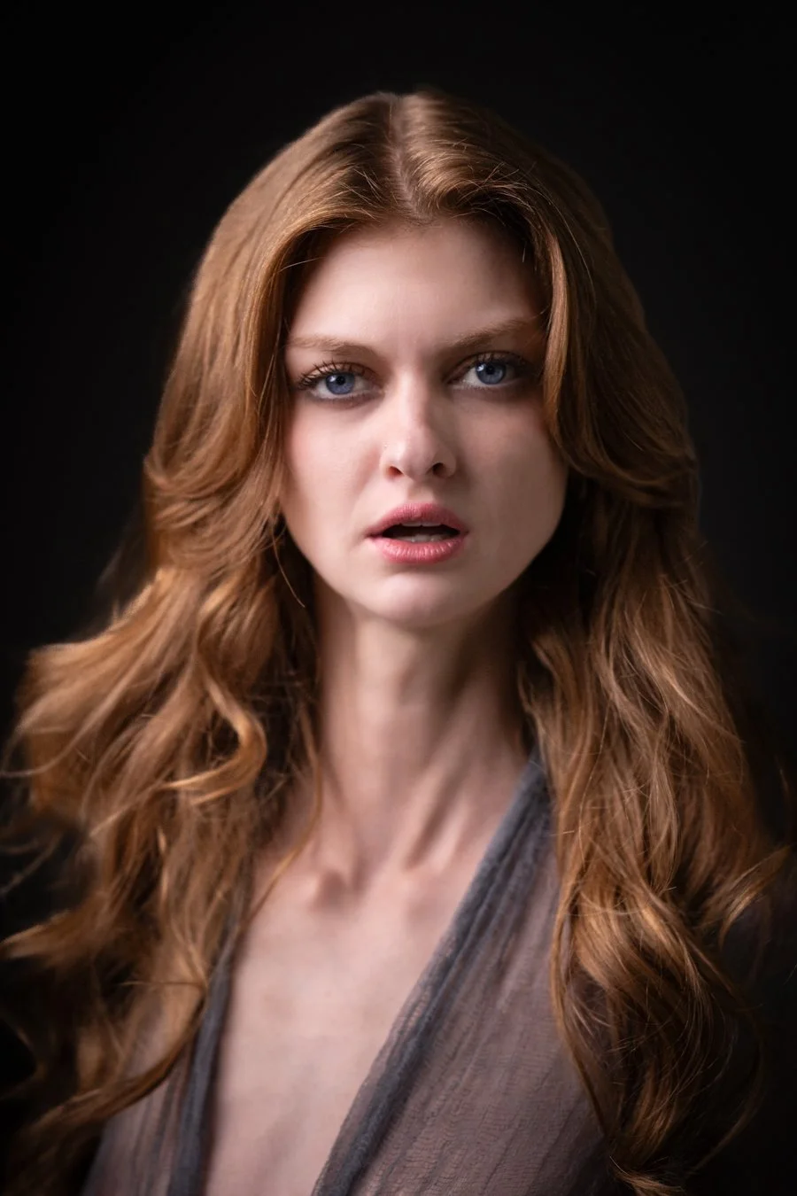 Close-up portrait of a woman with long, wavy red hair, blue eyes, and a surprised or confused expression, against a black background.