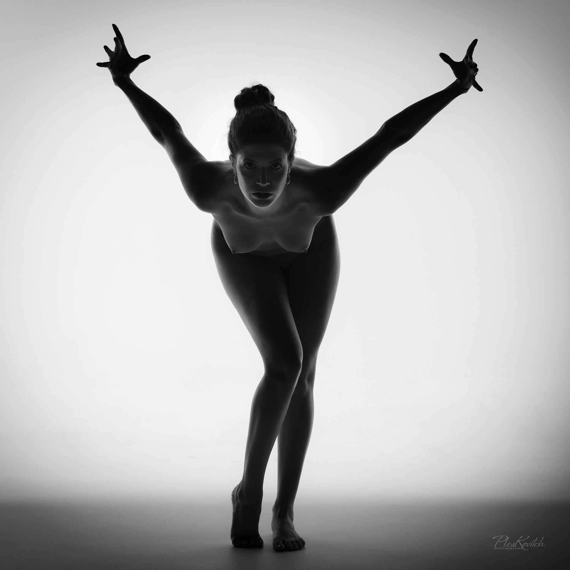 Black and white photo of a woman with her hair tied up, leaning forward with her arms extended to the sides, legs crossed, in a dynamic pose.