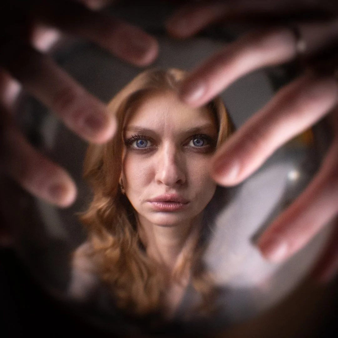 A woman with blue eyes and red hair looks into a mirror, with her hands framing her face.