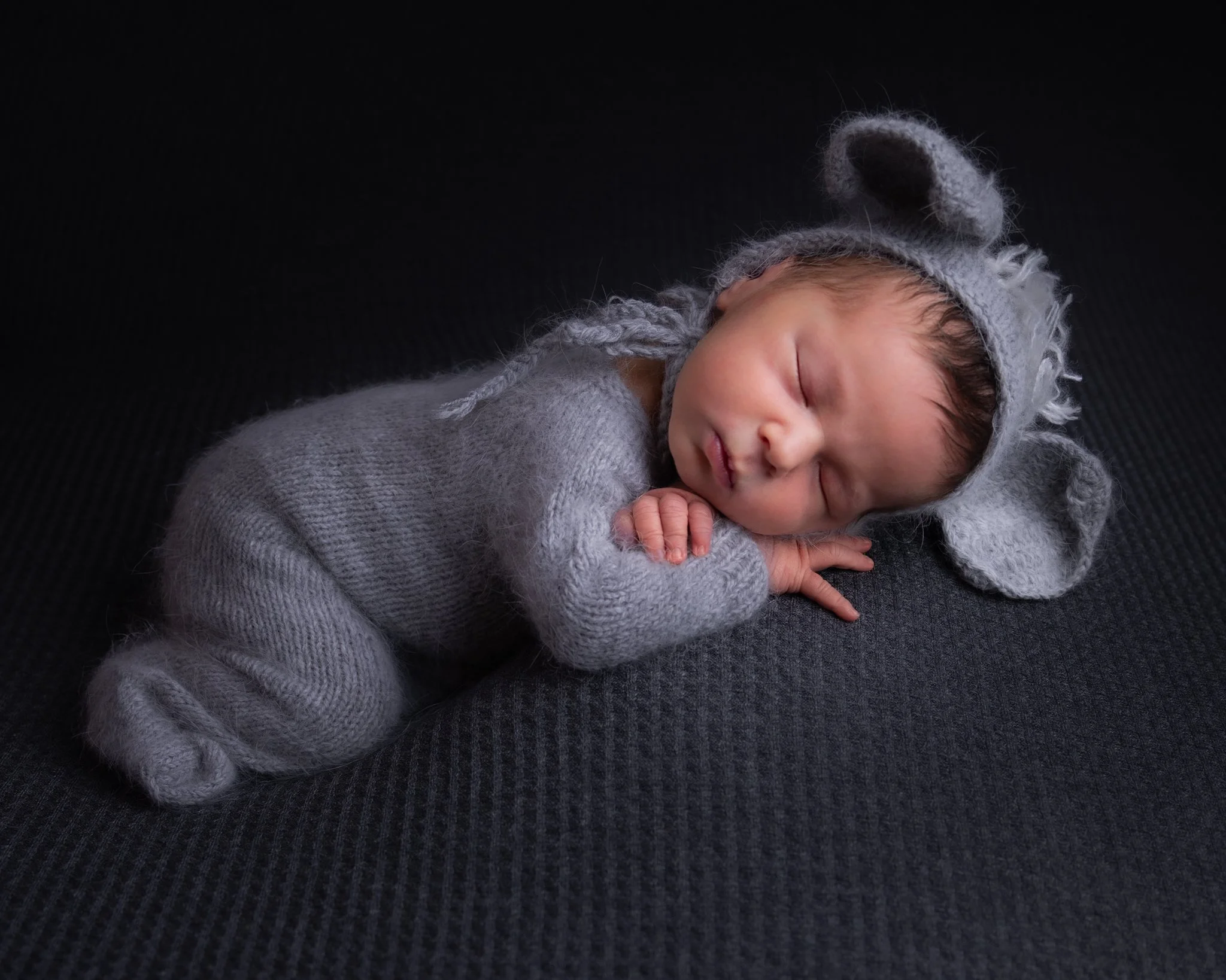 Close-up of a sleeping newborn baby wearing a gray knitted hat with ear and horn details, lying on a dark textured surface.