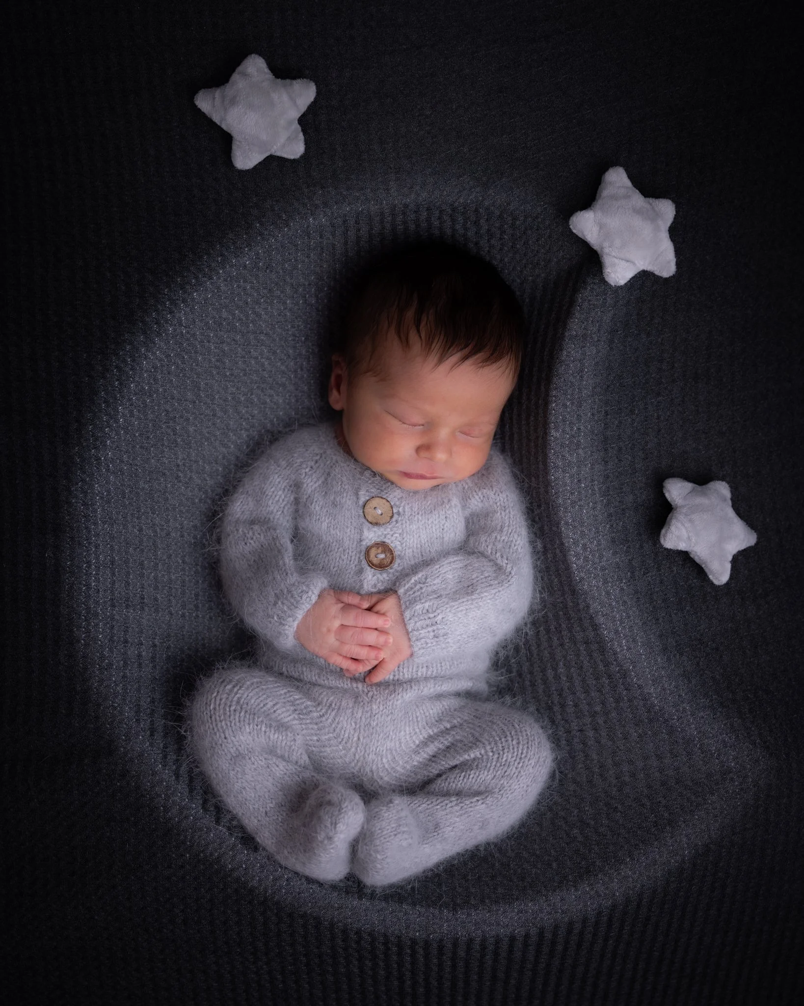 A sleeping baby with closed eyes, wearing a fuzzy gray sweater with wooden buttons, resting on a dark textured surface, surrounded by soft white star-shaped plush toys.