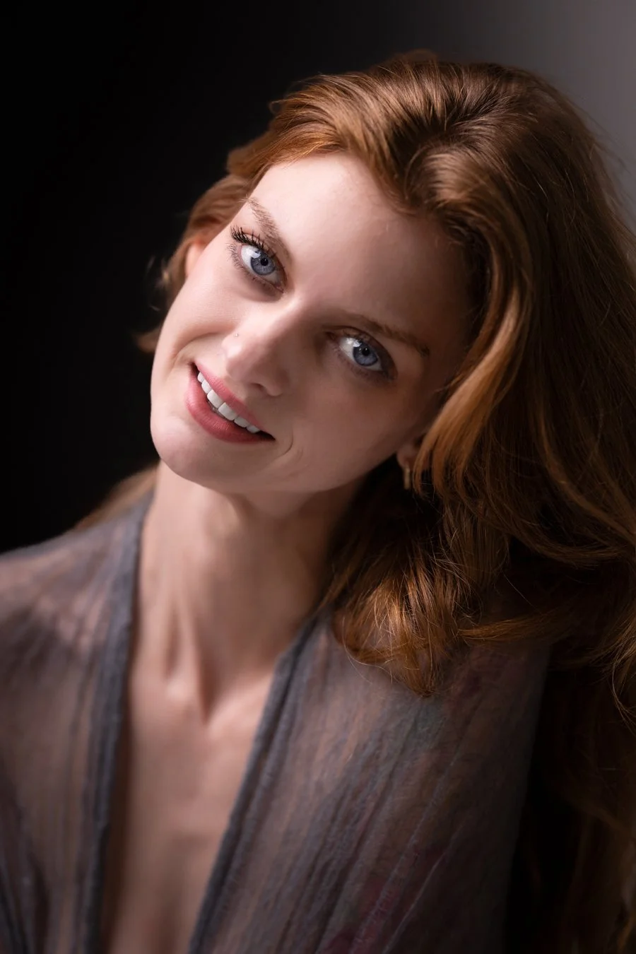 Close-up portrait of a woman with red hair and blue eyes, smiling slightly, wearing a sheer, striped top against a dark background.