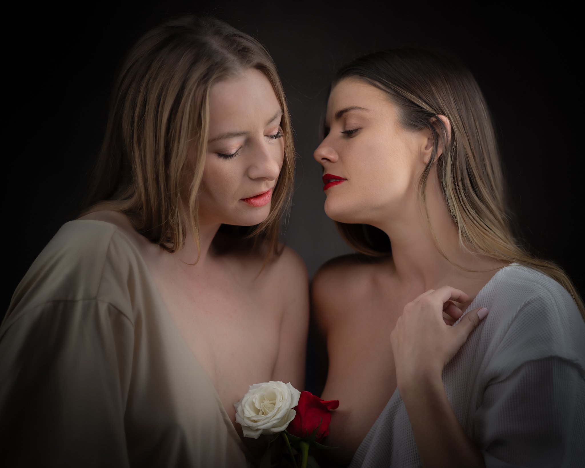 Two women sharing an intimate moment, their foreheads and noses touching, holding a bouquet of white and red roses, against a dark background.