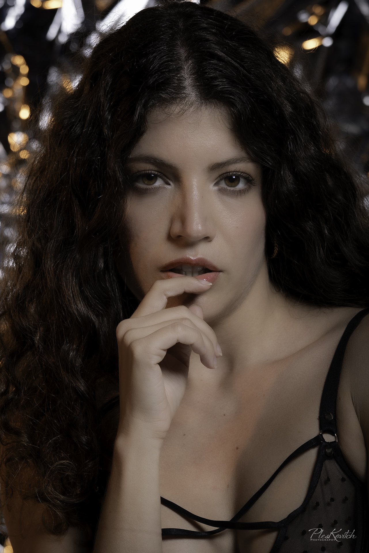 Close-up photograph of a woman with long, curly brown hair and hazel eyes, wearing black lingerie with sheer polka dot fabric, posing with her hand near her mouth. Blurred background with warm lighting.
