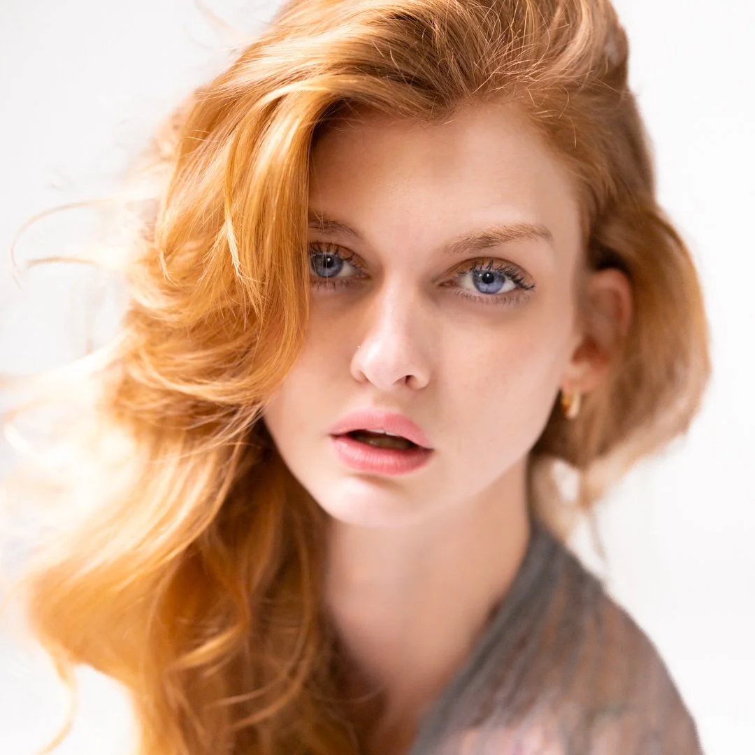 Close-up of a young woman with red hair and blue eyes, wearing a gray top, looking at the camera against a white background.
