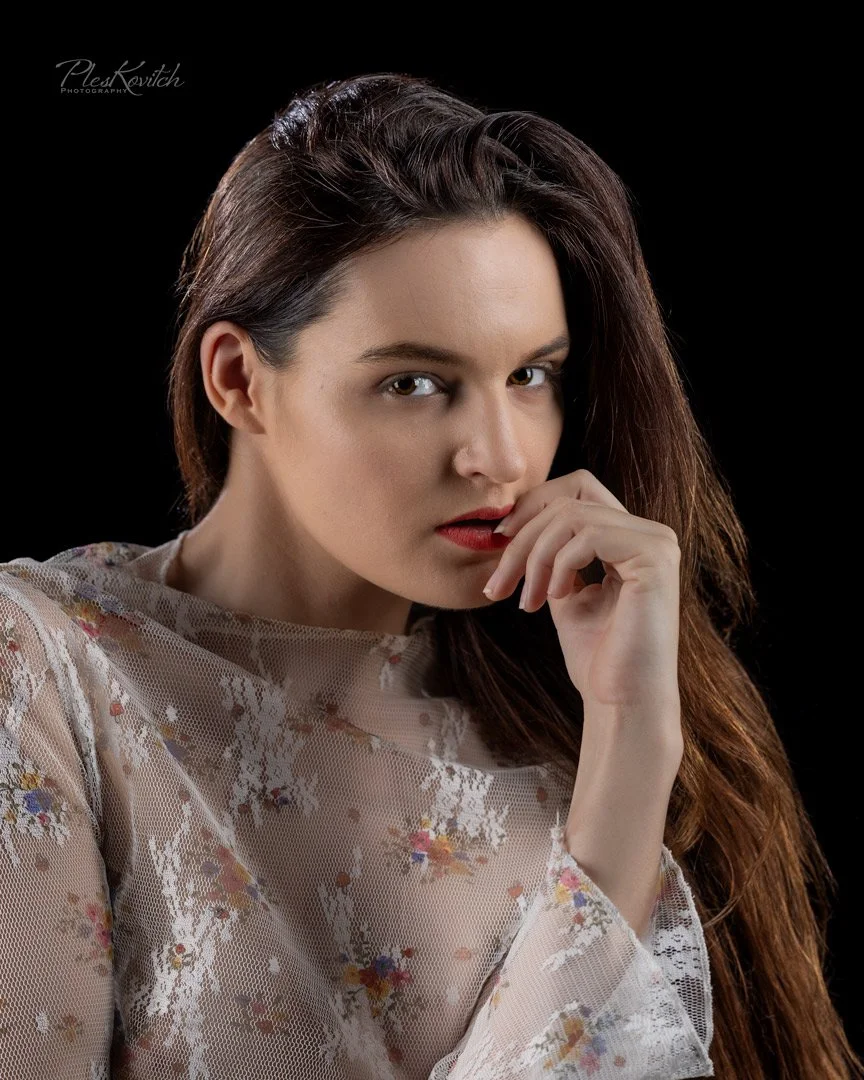 A woman with long brown hair, wearing a sheer lace top with floral patterns, posing with her hand near her face against a black background.
