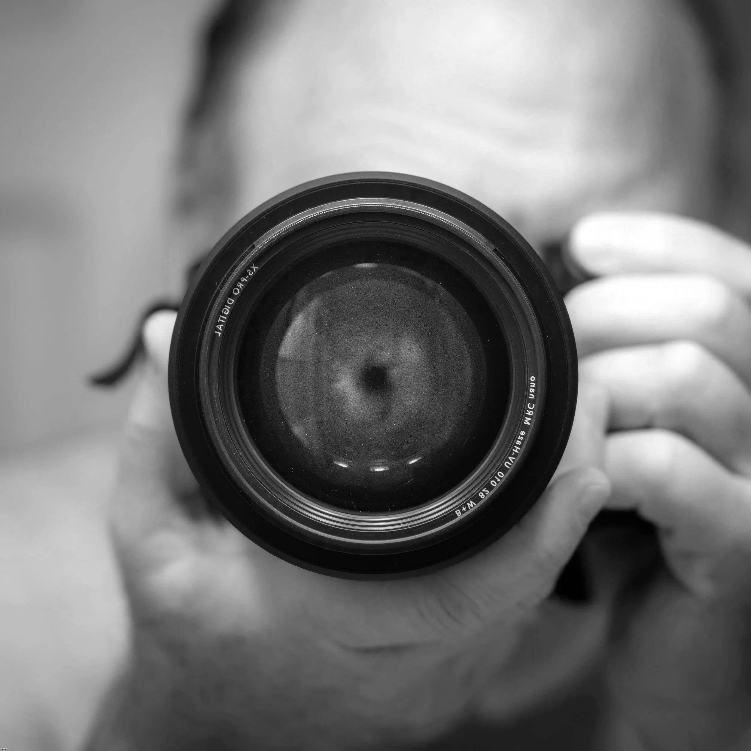 Close-up of a camera lens being held by a person, with the person's face blurred in the background, in black and white.