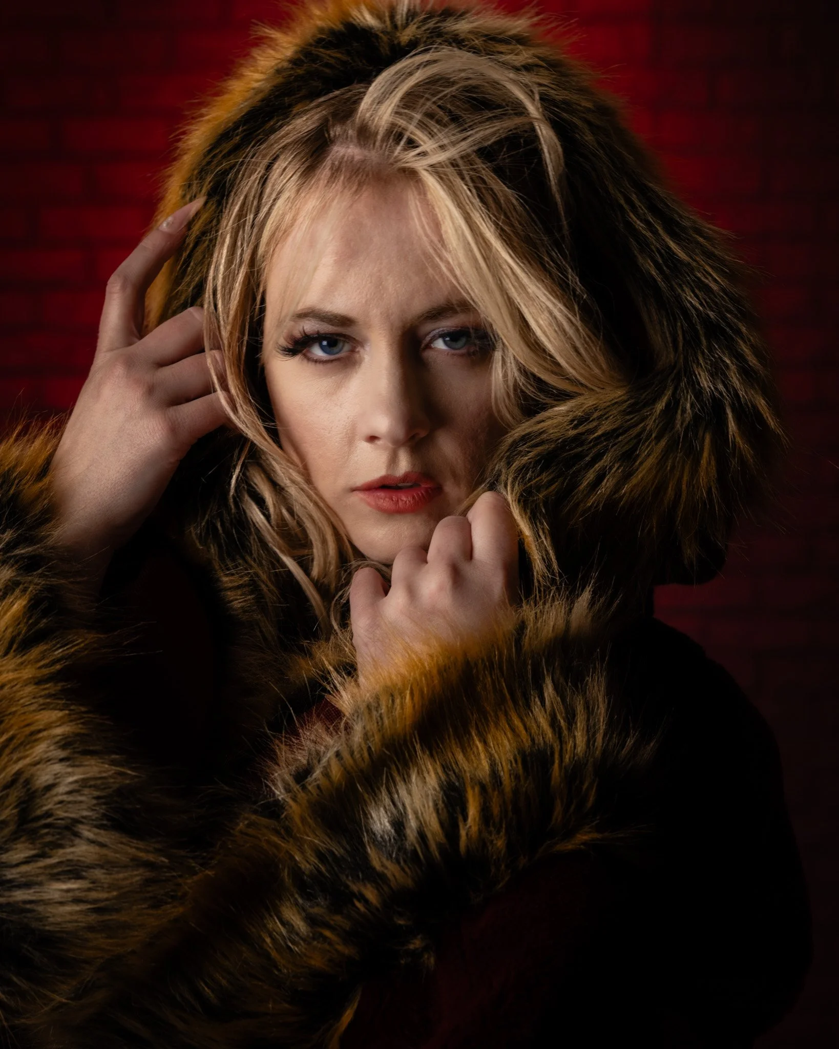 A woman with blonde hair and blue eyes wearing a fur coat, touching her head with one hand, against a dark red background.