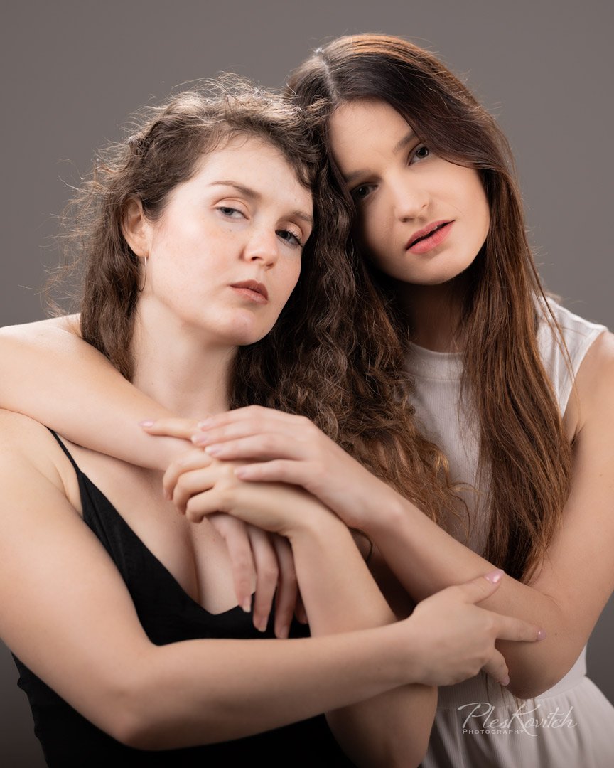 Two women are posing closely together, one with long wavy brown hair and the other with straight brown hair. They are touching and have neutral expressions, against a plain gray background.