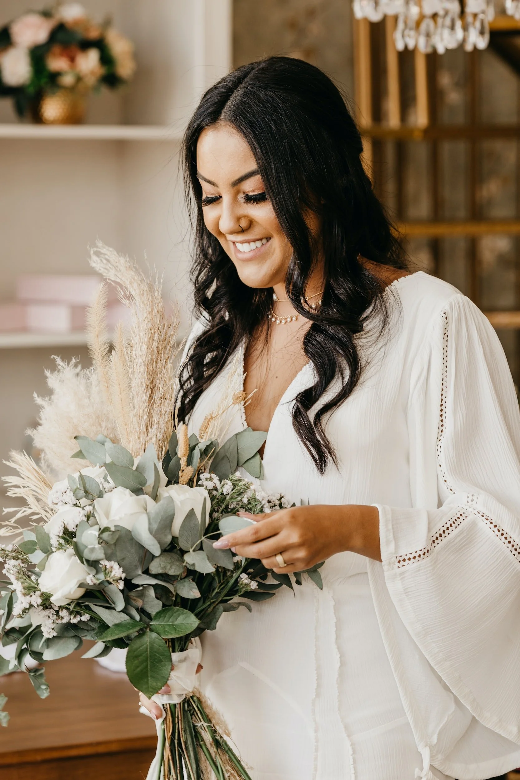 A woman with black wavy hair smiling and holding a bouquet of white roses, eucalyptus leaves, and pampas grass, wearing a white blouse with lace details and a pearl necklace.