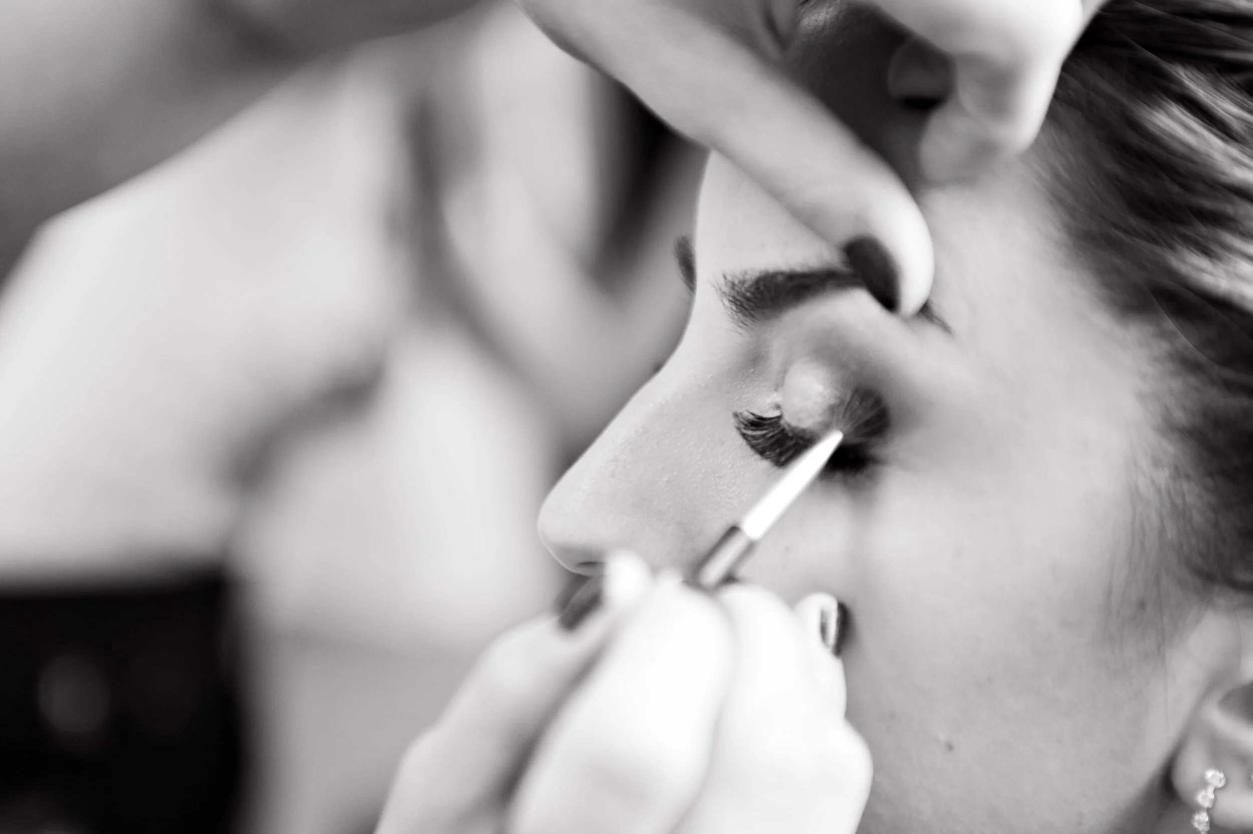 Close-up black and white photograph of a woman's face with her eyes closed, as makeup artist applies makeup using a brush.