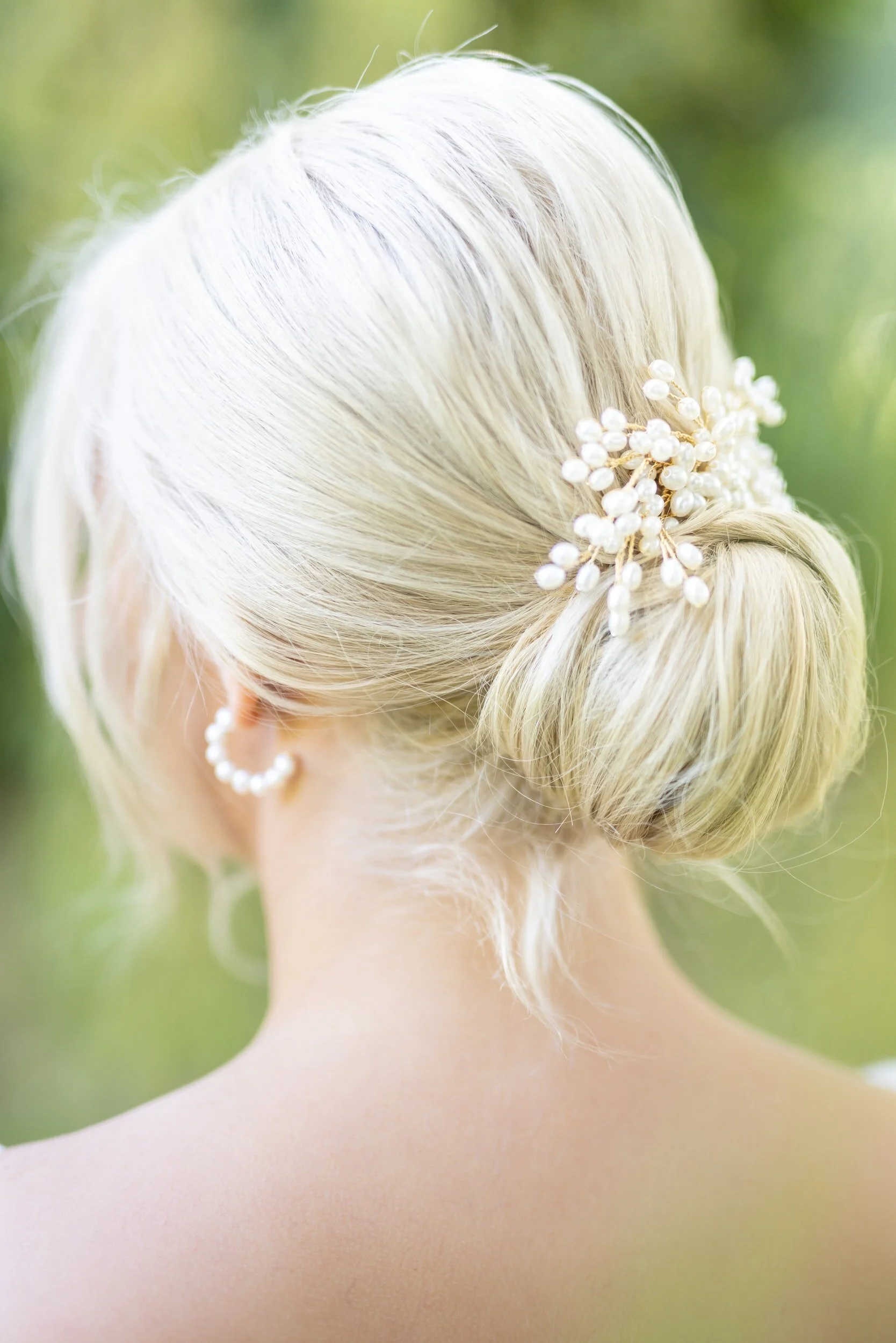 Back of a woman with platinum blonde hair styled in an elegant updo, adorned with a pearl hair accessory and wearing pearl earrings.