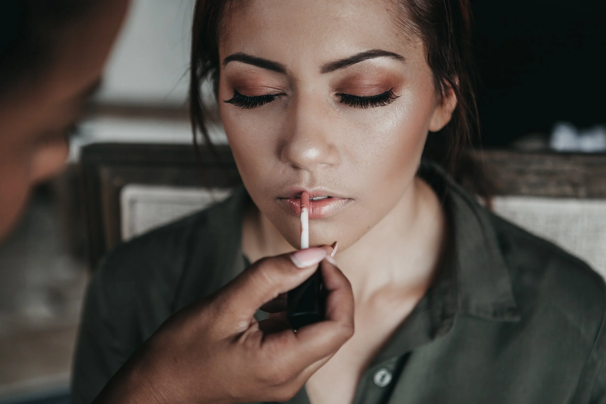 A woman with makeup and closed eyes having lipstick applied by another person.