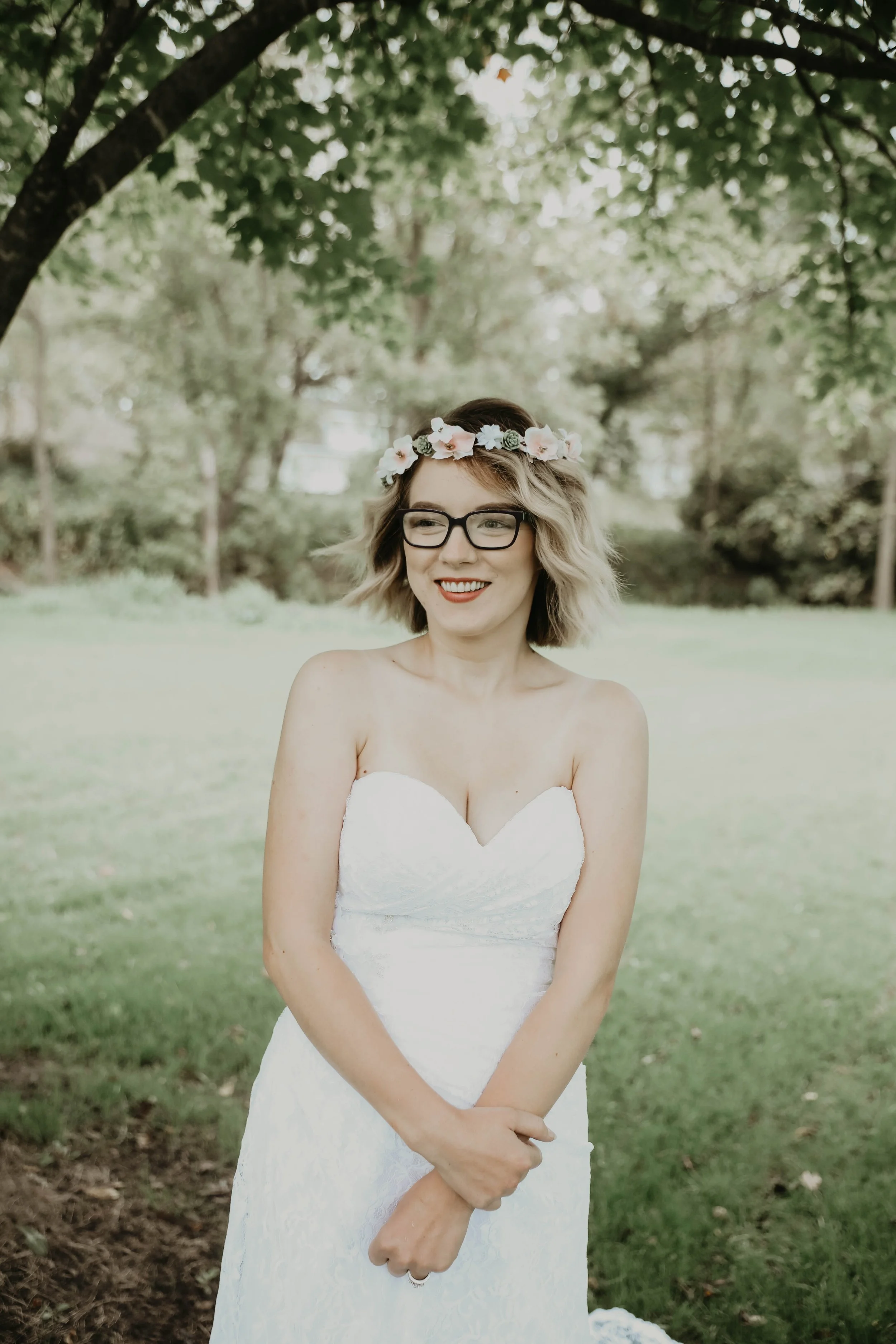 A smiling woman wearing glasses and a flower crown, dressed in a white strapless wedding gown, standing outdoors on a grassy area with trees in the background.