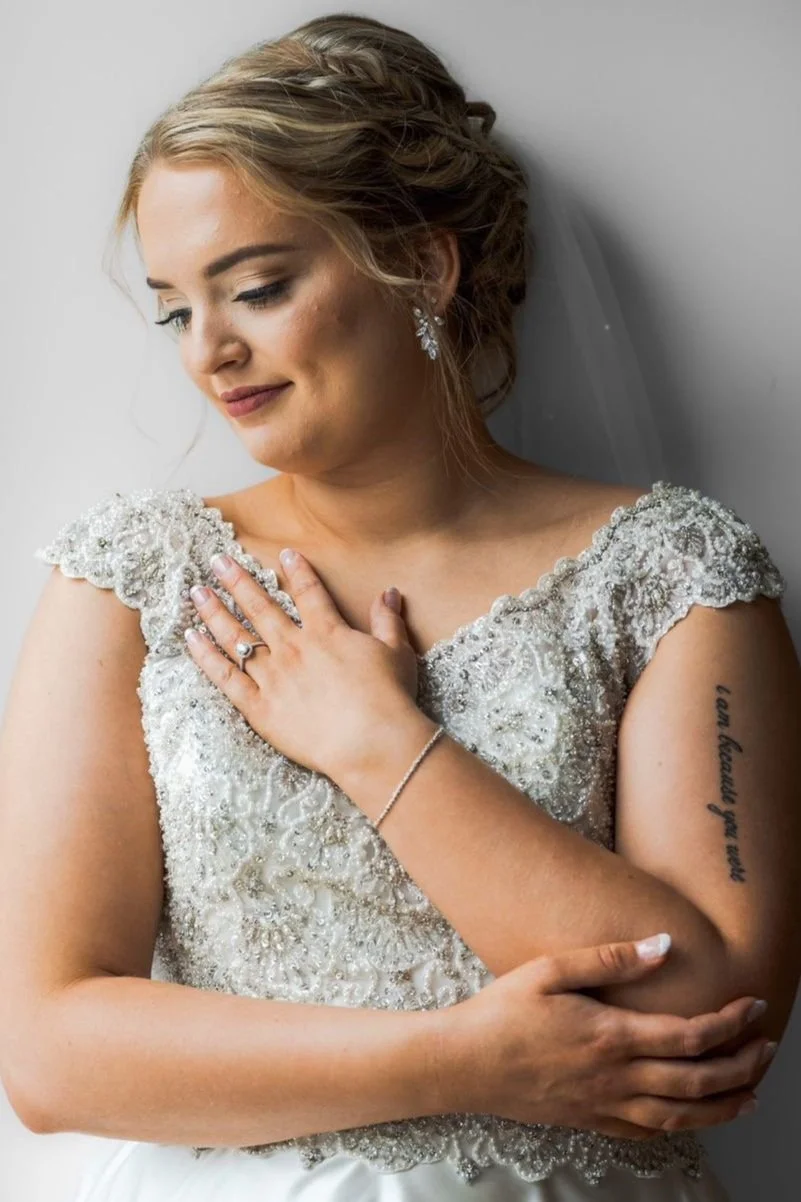 A bride with blonde hair styled in an updo, wearing a detailed, beaded wedding dress and earrings, standing against a light background, with her eyes closed and one hand resting on her chest, showing a wedding ring and a tattoo on her arm.