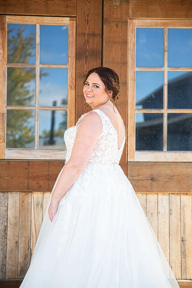 A smiling bride in a white wedding dress standing outdoors in front of a rustic wooden building with large windowpanes reflecting the sky and trees.