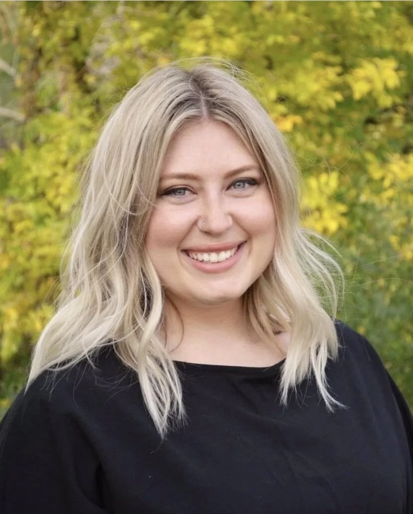 A young woman with blonde hair and a nose ring smiling outdoors with green and yellow foliage in the background.