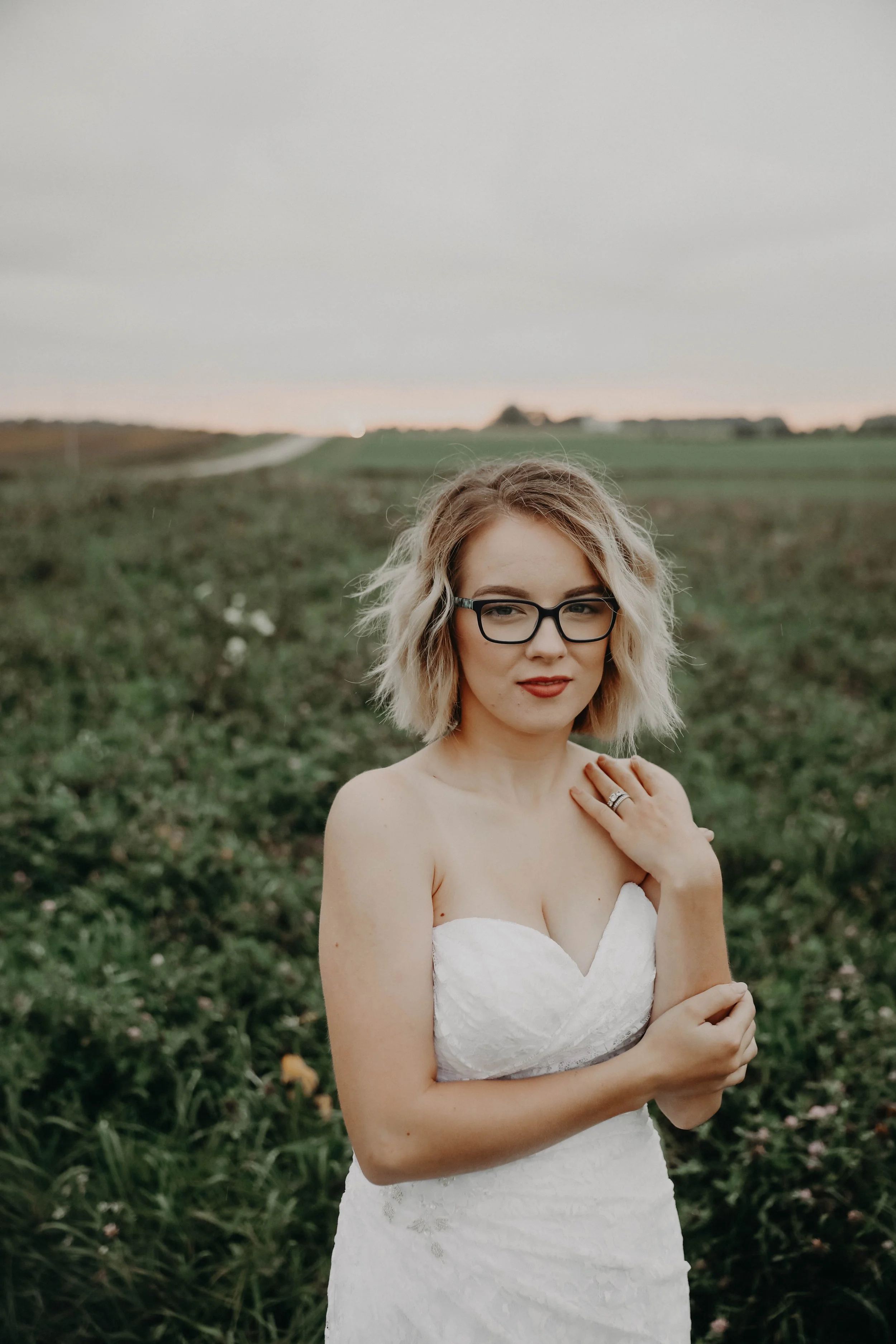 A woman with blonde, curly hair wearing glasses and a white strapless dress standing outdoors in a field at sunset.