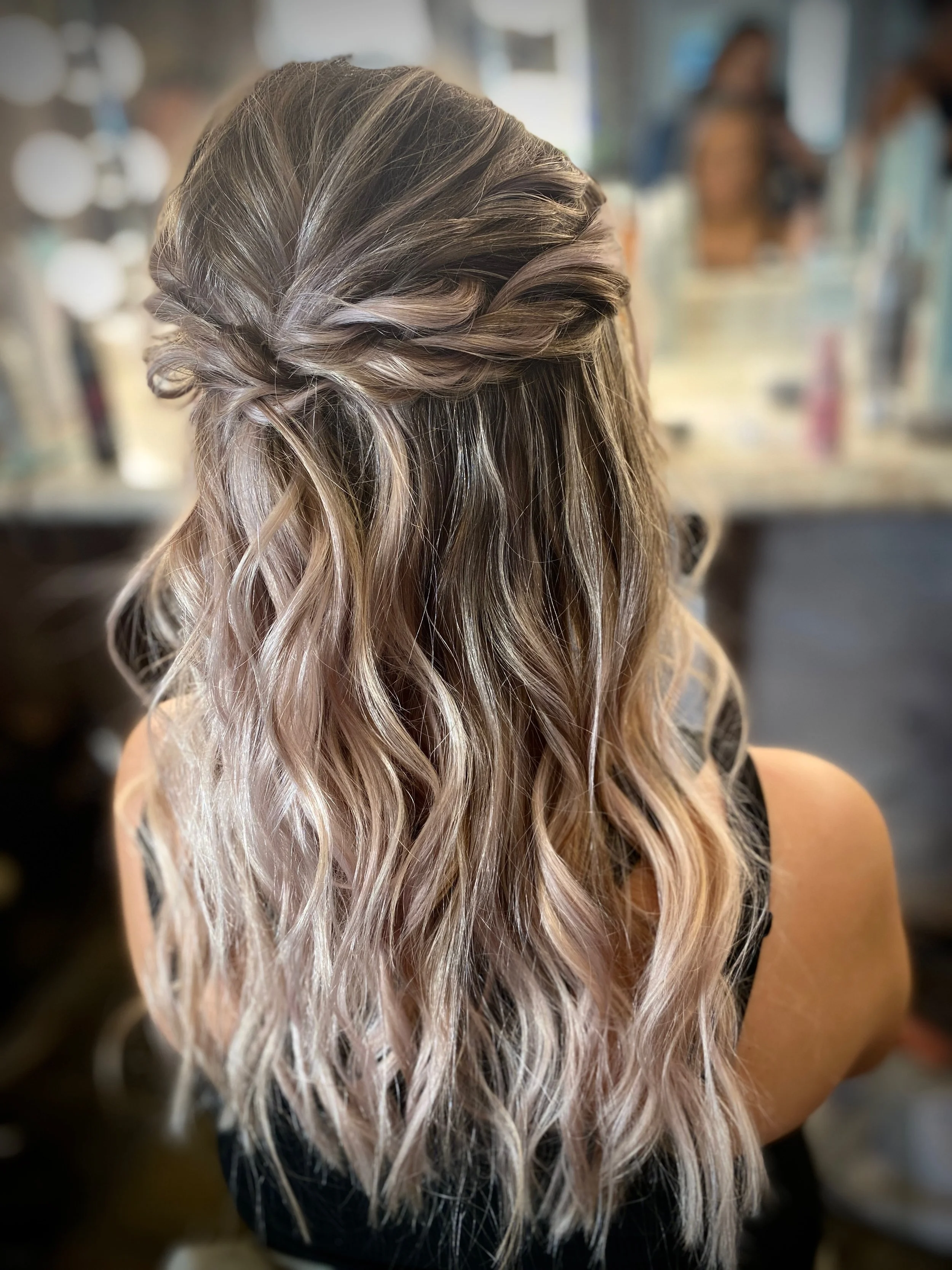 Back view of a woman with long, wavy, blonde hair styled in loose curls with a twist at the top, sitting at a salon or similar setting.