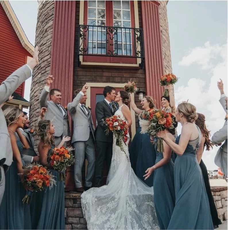 A bride and groom kissing on their wedding day surrounded by bridesmaids and groomsmen outside a building with a stone and red siding facade, with some of the guests raising their hands in celebration.