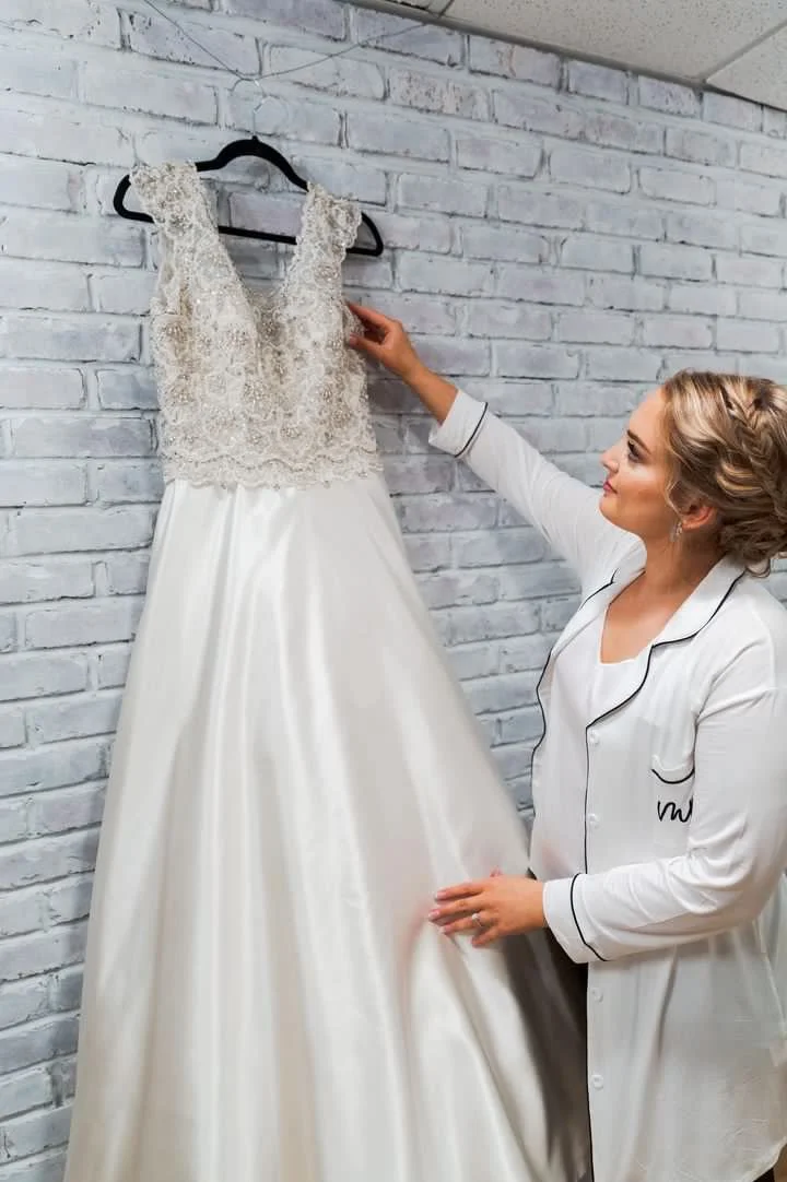 A woman in pajamas holding a wedding dress hanging on a black hanger against a white brick wall.