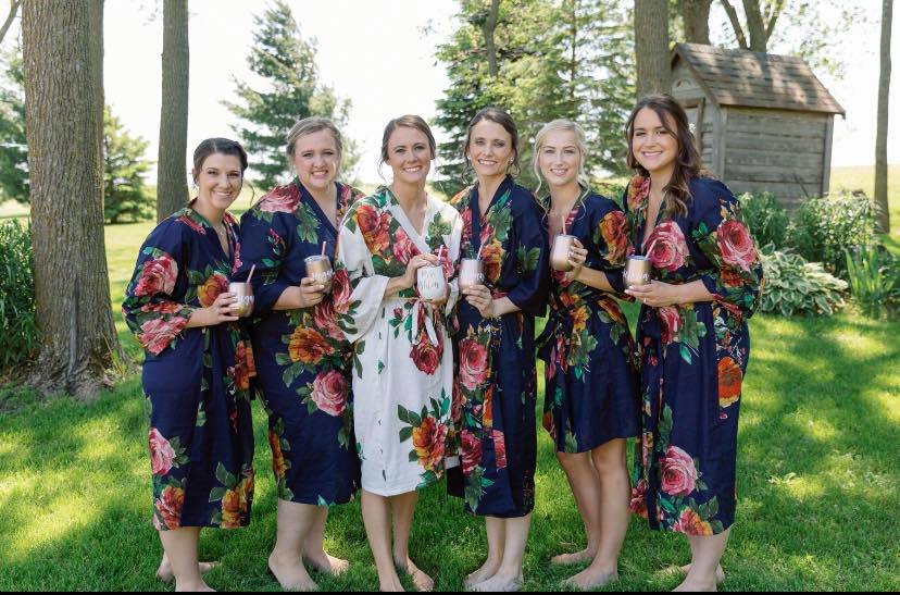 Six women in floral robes standing outdoors on grass, holding drinks, with trees and a wooden shed in the background.