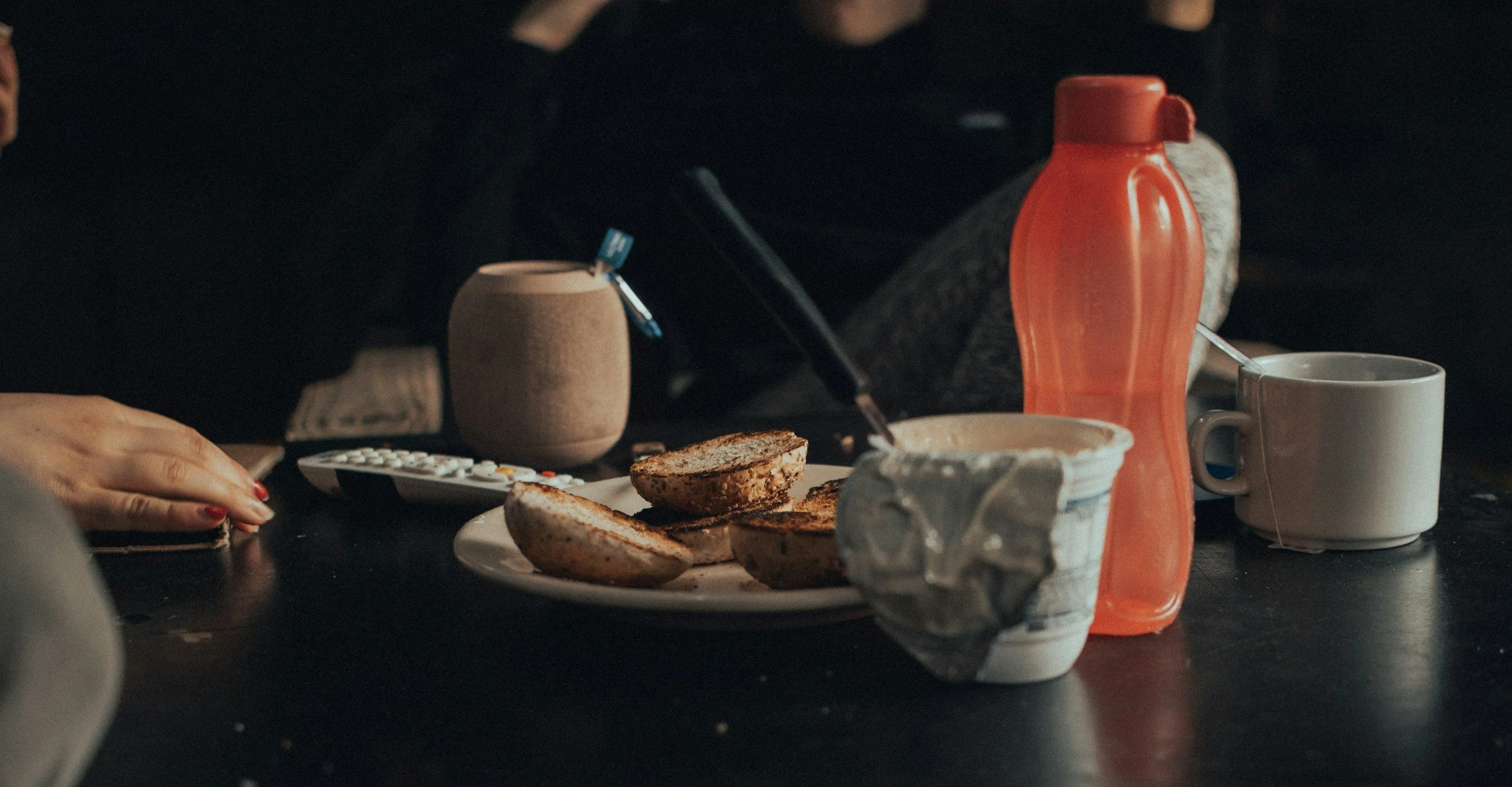 A breakfast table with toasted bread, a mug, a vibrant orange water bottle, and a small container, with a hand resting nearby and a remote control on the table.