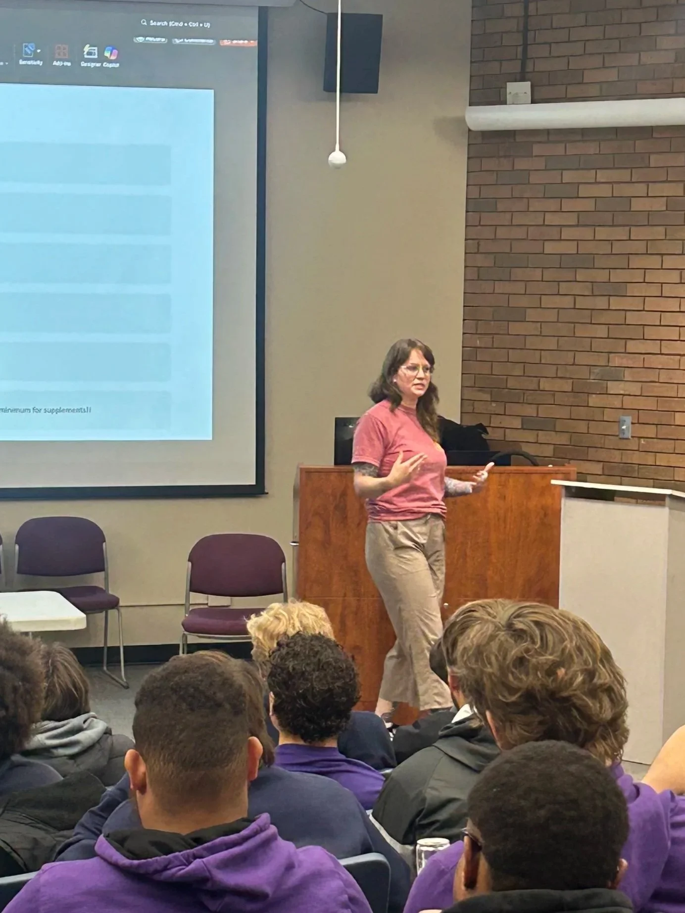 A woman giving a presentation in a classroom or lecture hall. She is standing near a large screen that displays a computer interface, with an audience of students, some wearing purple hoodies, listening attentively.
