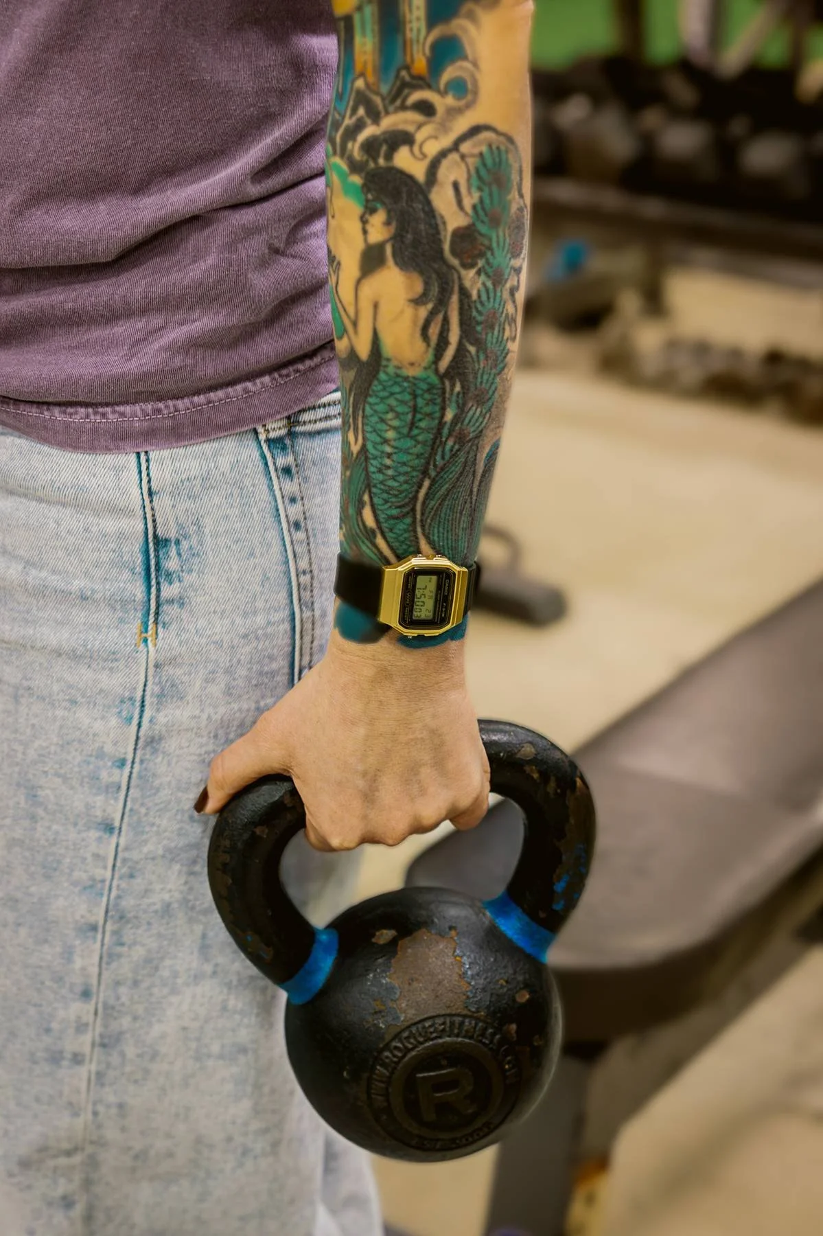 A person with colorful tattoos on their arm, wearing a purple shirt and light blue jeans, holding a black kettlebell with weathered paint, in a gym setting.