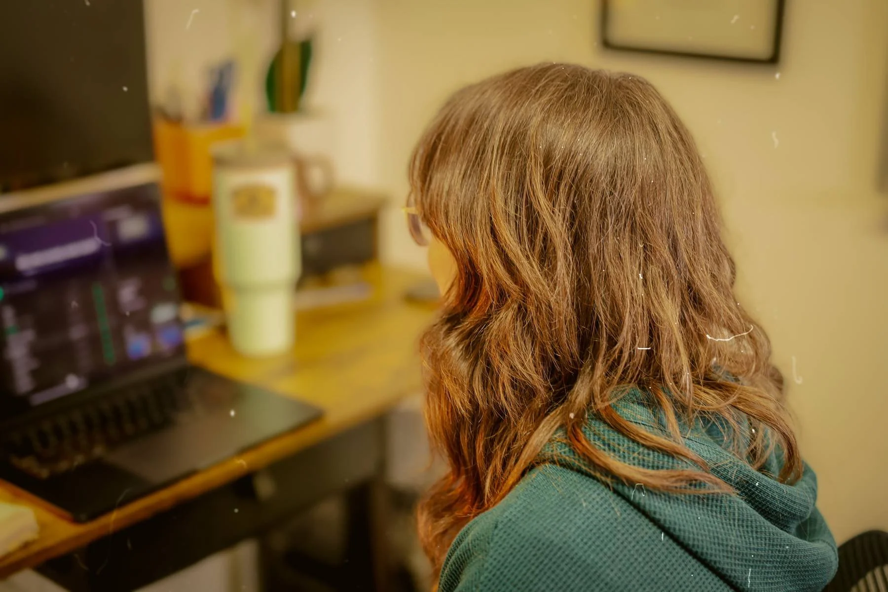 A woman with wavy brown hair wearing glasses and a green sweater sitting at a desk with a computer, books, and a yellow mug in a cozy room.