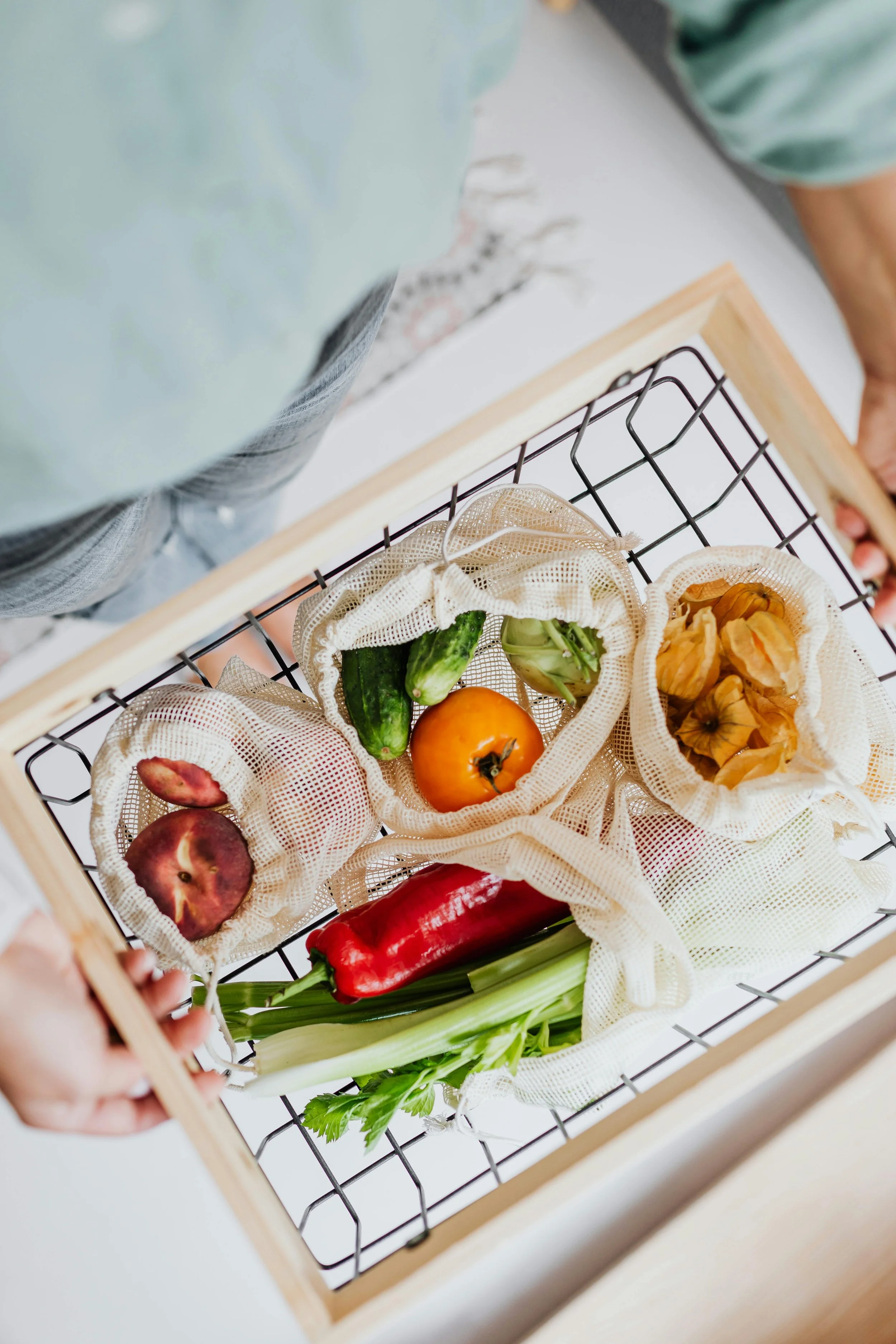 Basket containing fresh produce including a peach, a tomato, cucumbers, green onion, a red pepper, and dried flower petals.