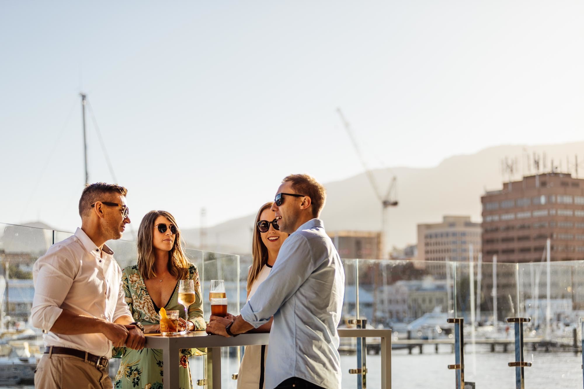 Group of four people socializing outdoors with drinks, against a backdrop of a marina and cityscape.