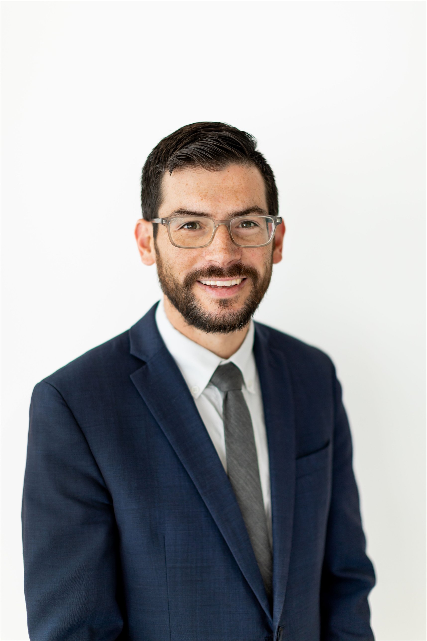 A professional man with glasses and a beard wearing a navy blue suit, white shirt, and gray tie, smiling against a white background.