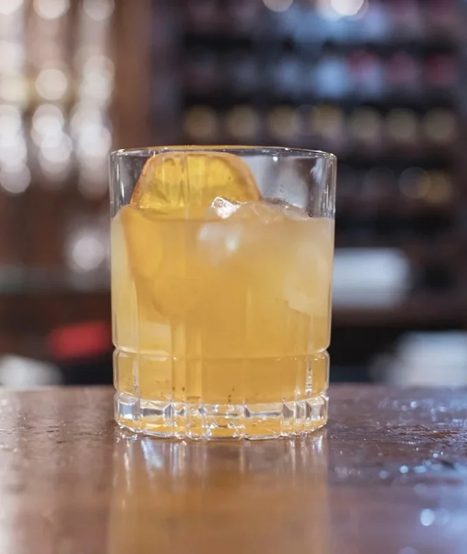 An Old Fashioned cocktail, in a glass with ice and a lemon wedge on a wooden bar counter.