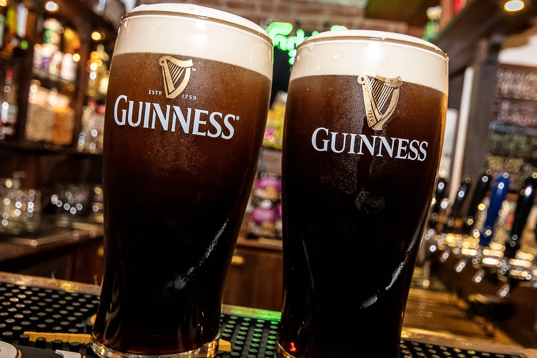 Two pints of Guinness beer with foam on top, on a bar counter, with blurred background of a bar setting and illuminated sign.