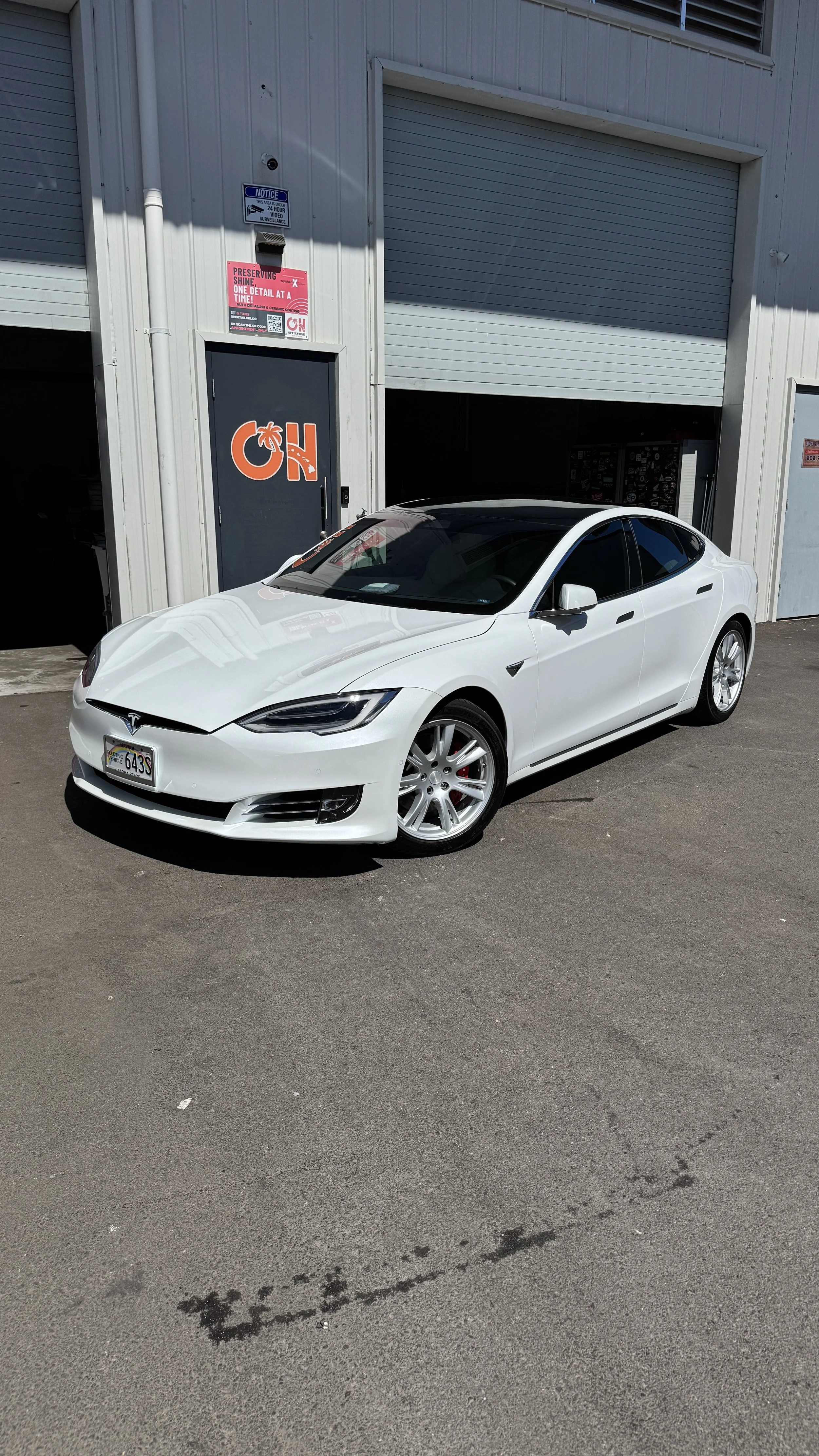 White Tesla Model S parked outside a commercial building with a gray metal exterior and an orange 'OH' sign on the door.