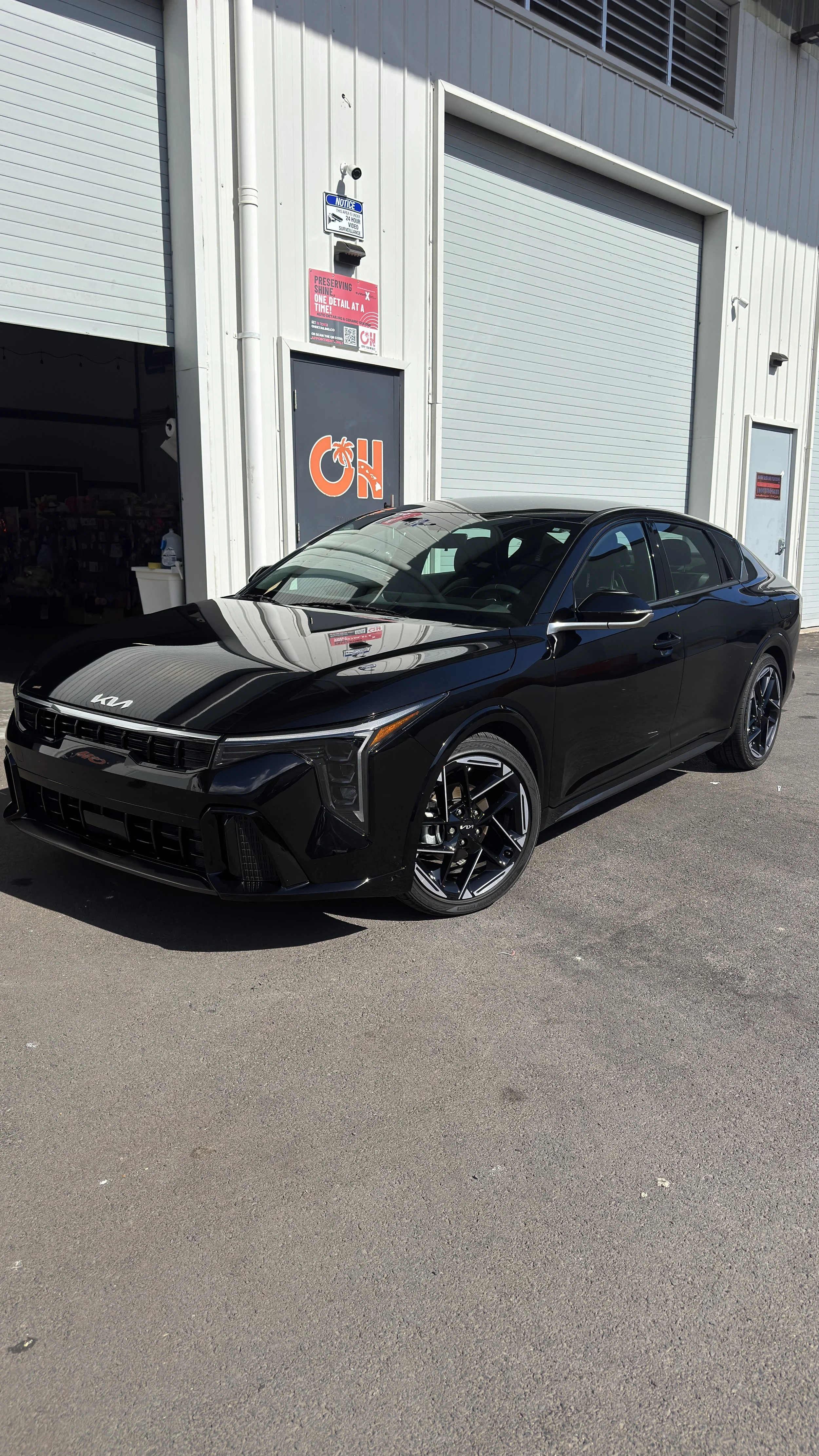 A black Kia sedan parked outside an industrial building with a gray metal exterior, a raised garage door, and a sign with the letters 'OH' on the wall.