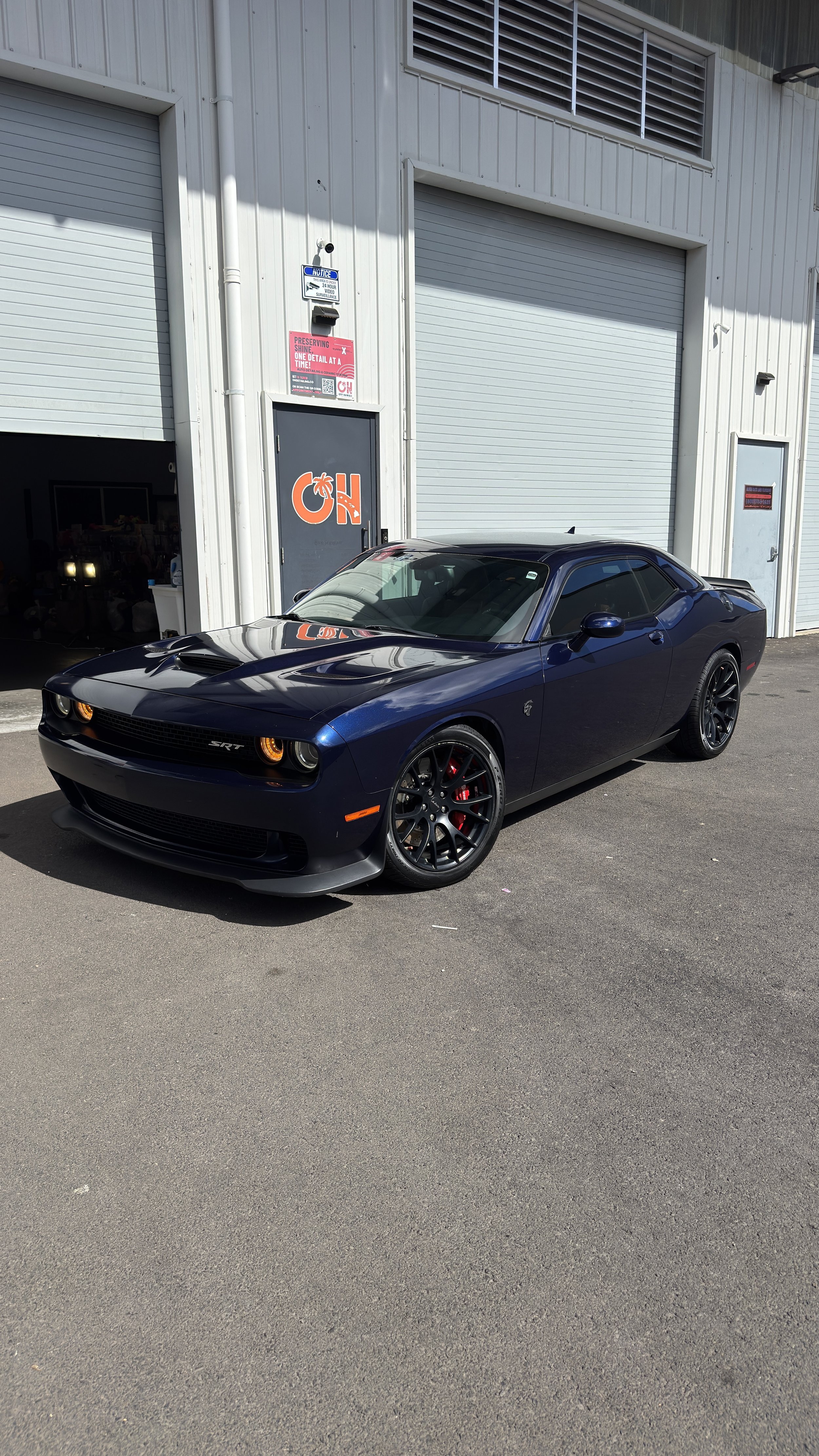 A black Dodge Challenger SRT parked outside a commercial building with large gray metal garage doors and a sign with the letters 'OH' in orange.